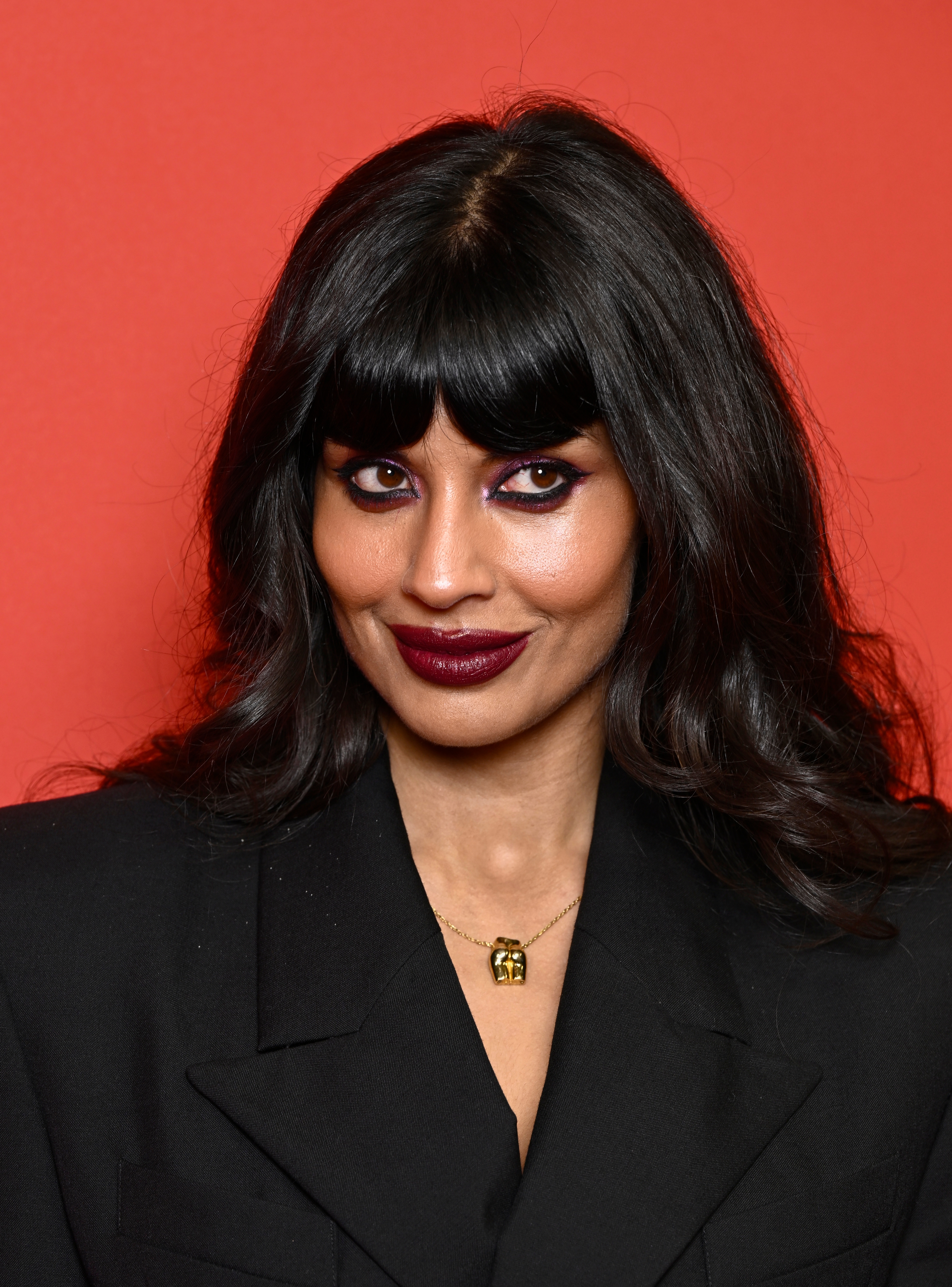 Jameela Jamil with agelong  hair, wearing stylish blazer and elephant necklace, smiles somewhat  against a plain backdrop