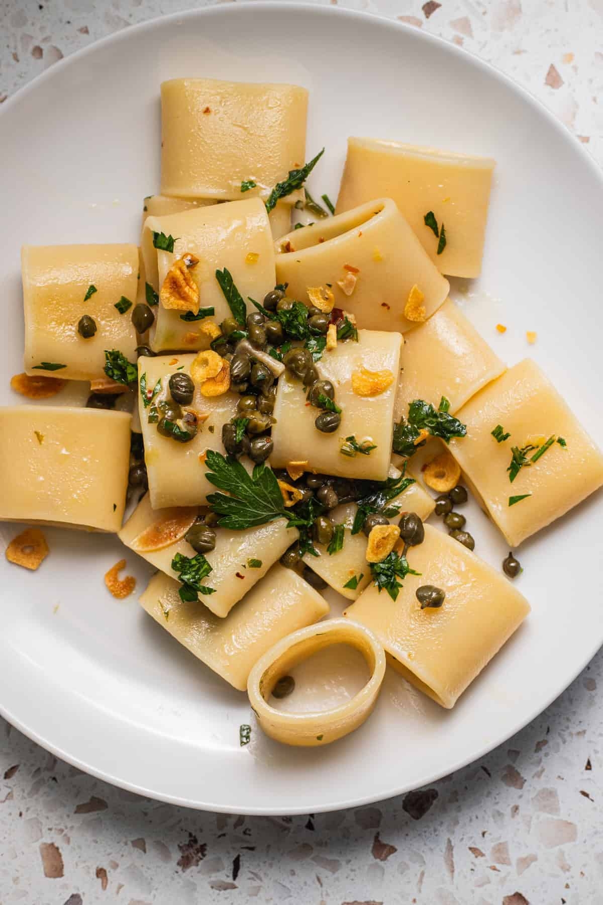 A sheet of paccheri pasta with capers, ail slices, and caller parsley, served connected a achromatic dish