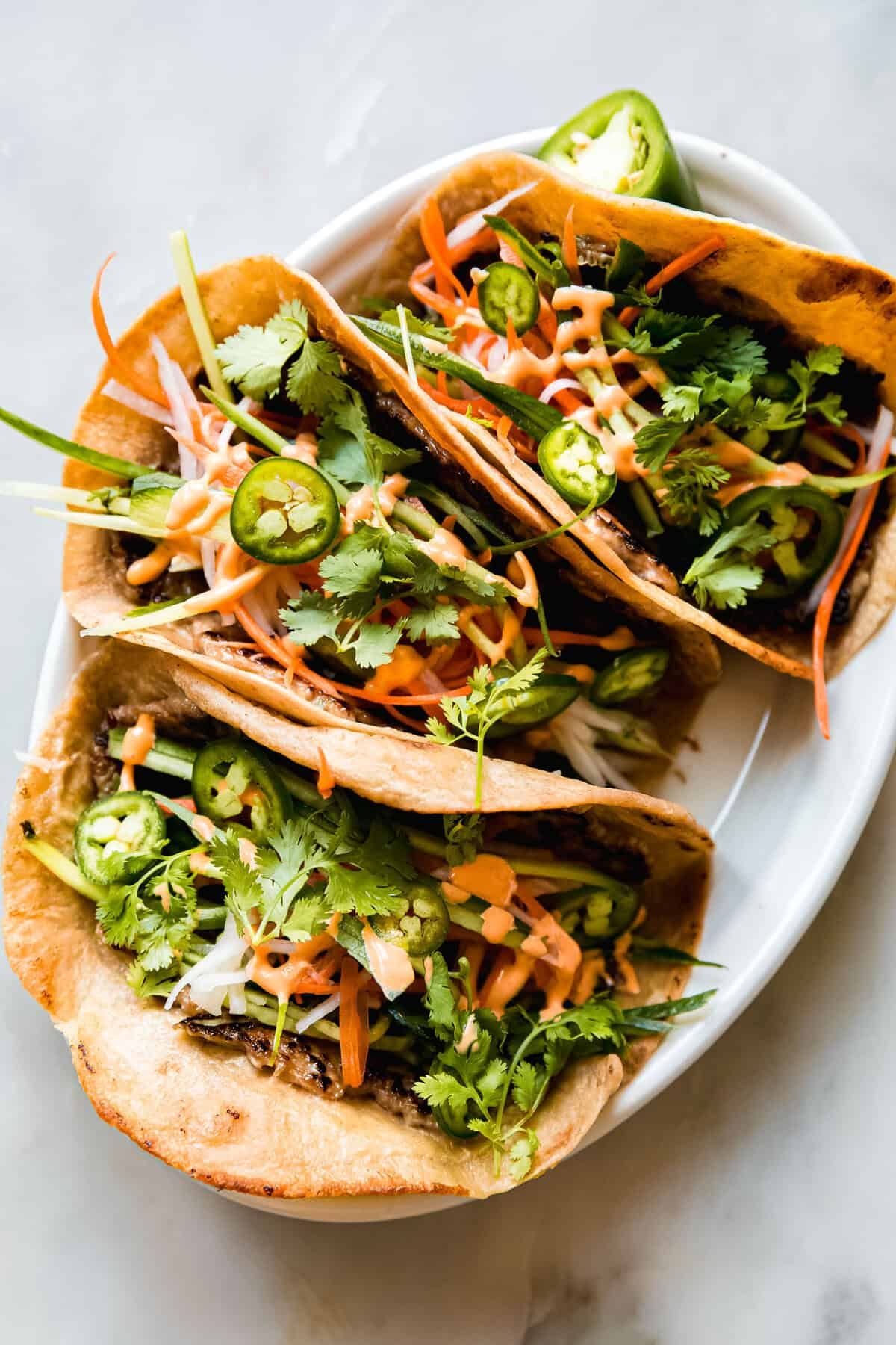 Three tacos connected a plate, filled with vegetables similar cilantro, sliced jalapeños, carrot shreds, and drizzled with a spicy sauce
