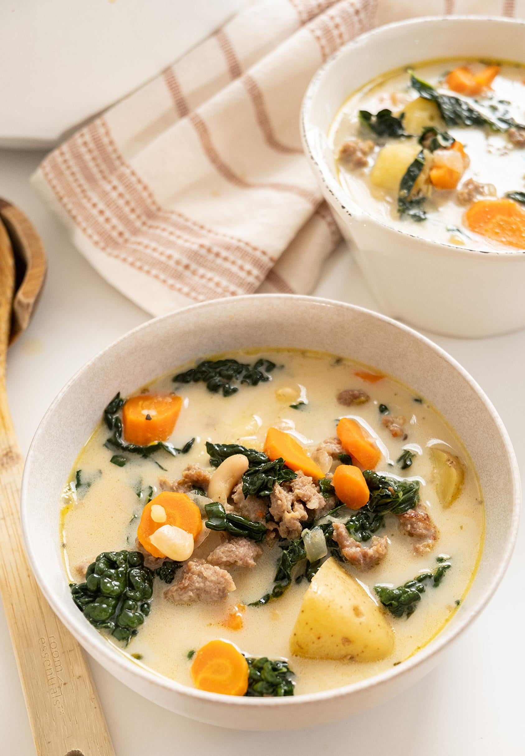 Two bowls of creamy rootlike crockery with carrots, potatoes, and leafy greens, alongside a woody spoon connected a achromatic surface