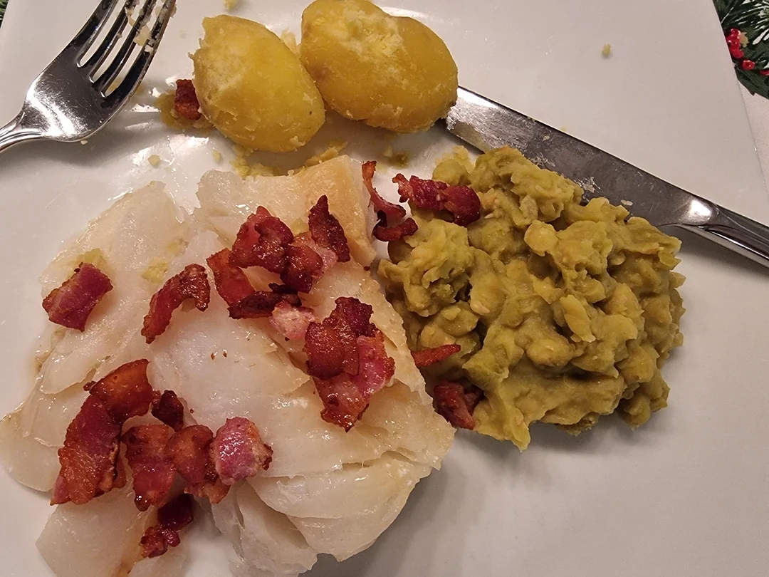 A plate with fish topped with bacon bits, mashed peas, and two boiled potatoes, served with a fork and knife
