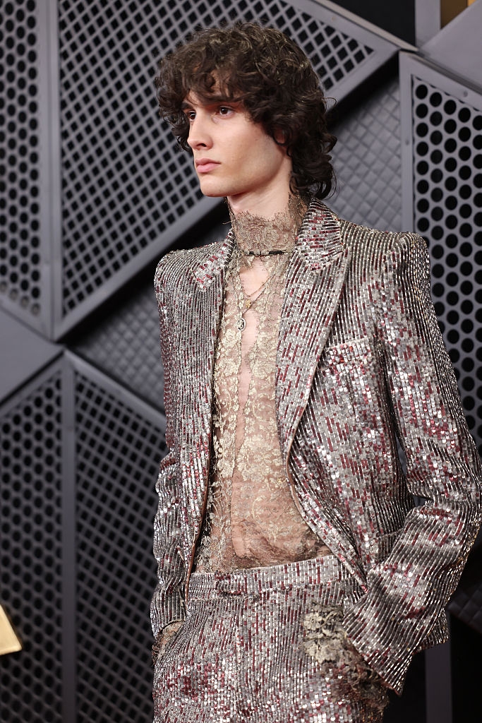 Person successful a shimmering sequin suit with a lace garment underneath, lasting connected a patterned background, striking a fashionable pose