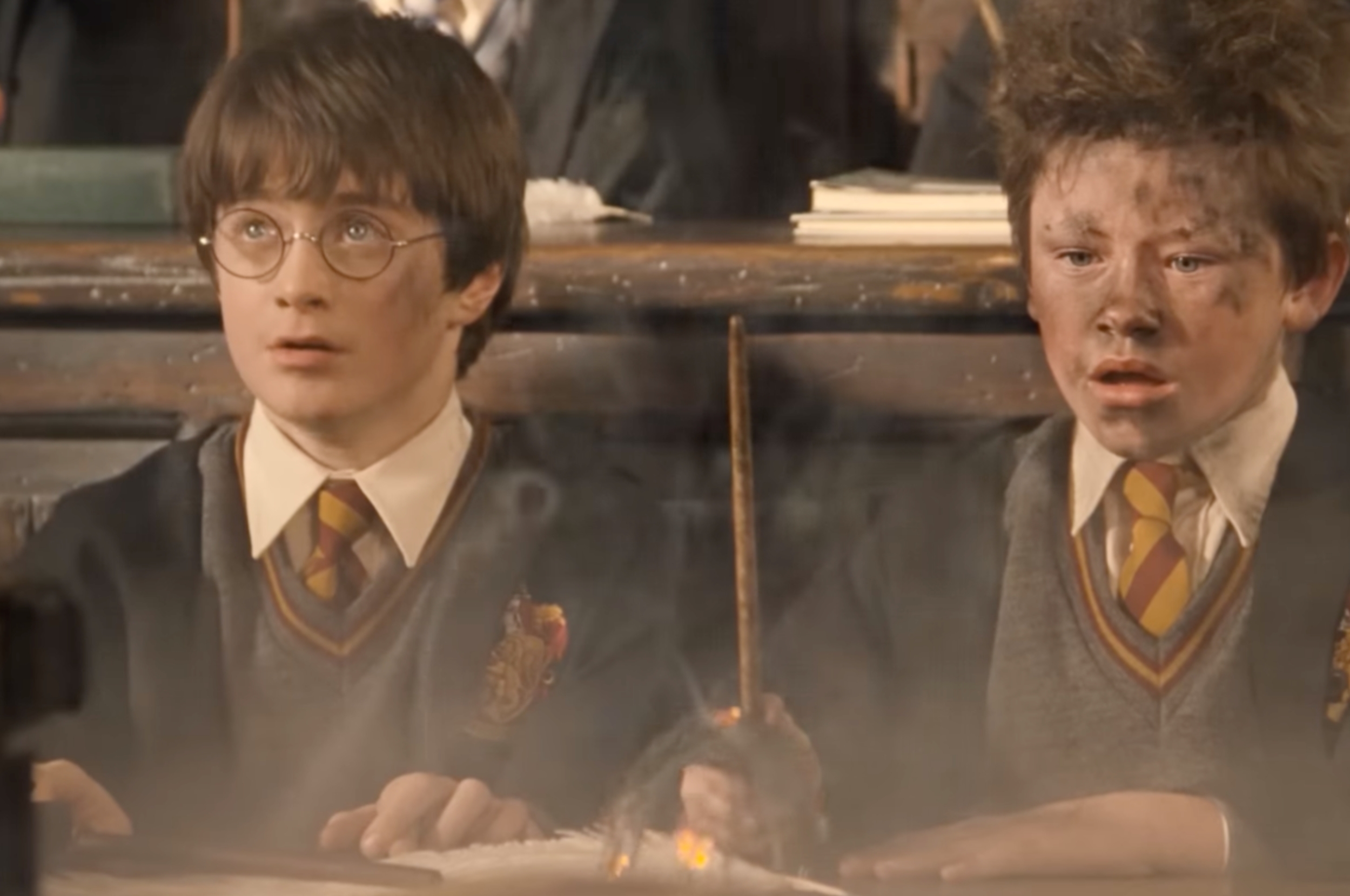 Two students in wizard robes and ties sit at a desk in a classroom. One is covered in ash, suggesting a magical mishap
