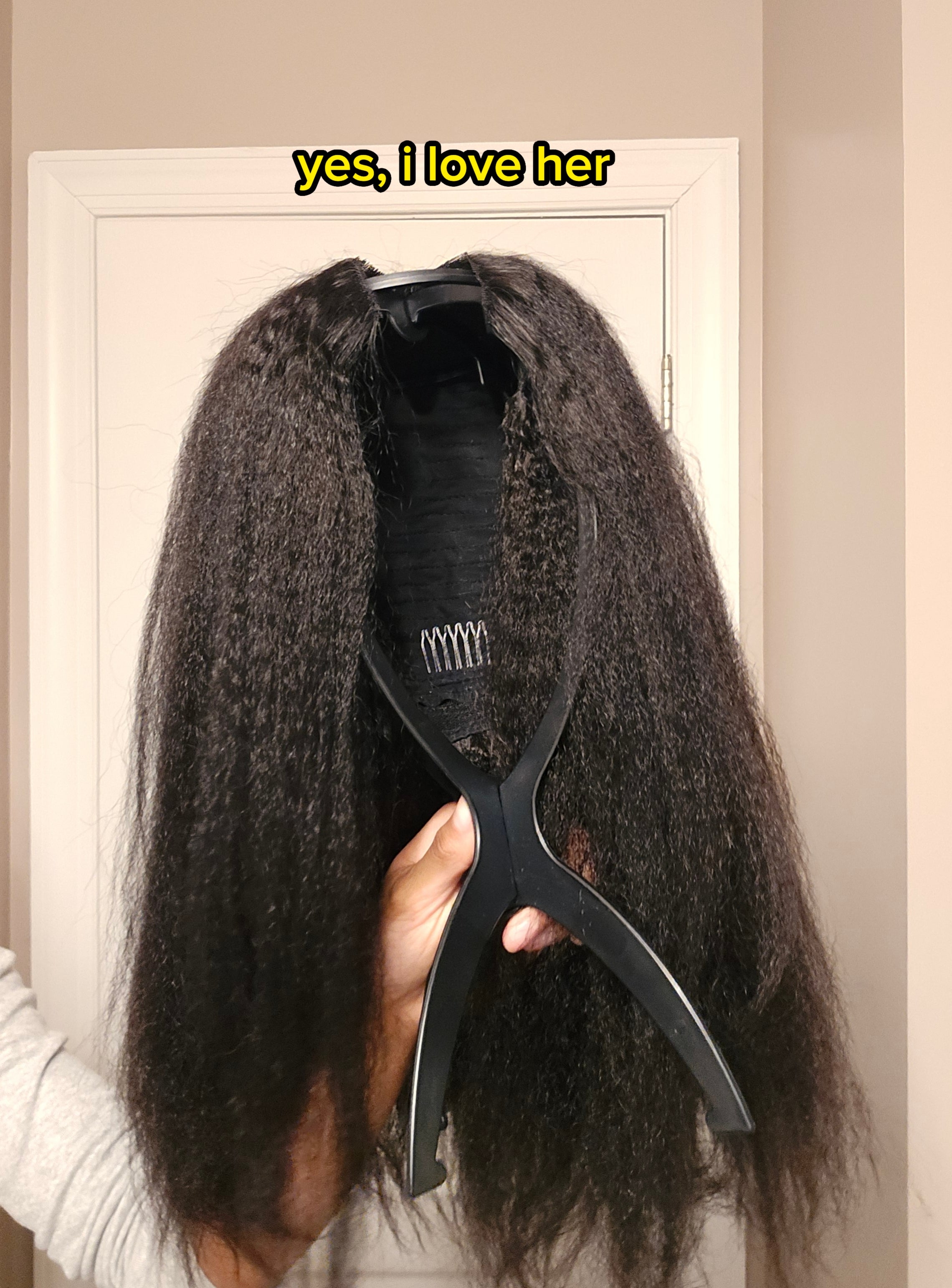 A wig held up by a stand, featuring long, textured hair, displayed indoors against a plain doorway background
