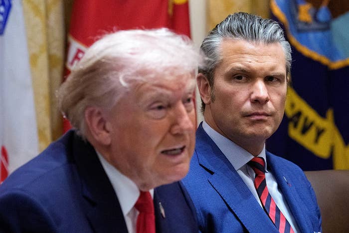 Two men are seated next to each other at a table. The man closer to the camera is speaking, while the other listens attentively in a formal setting