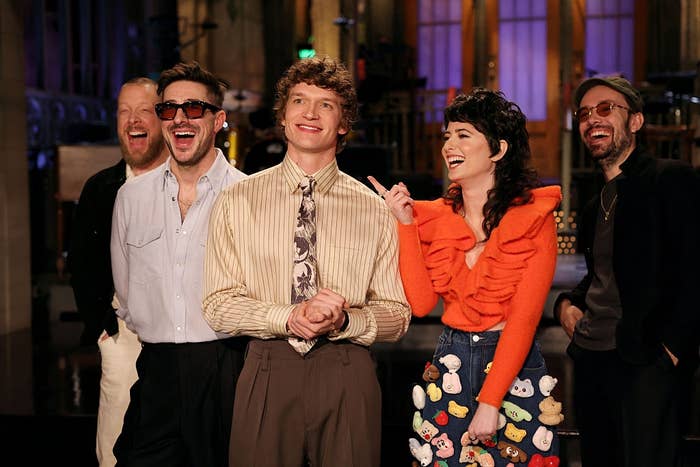 Group of 5  radical   laughing unneurotic  indoors, 1  wearing a ruffled apical  with cartoon patches. They're connected  a workplace  set, perchance  for a TV show