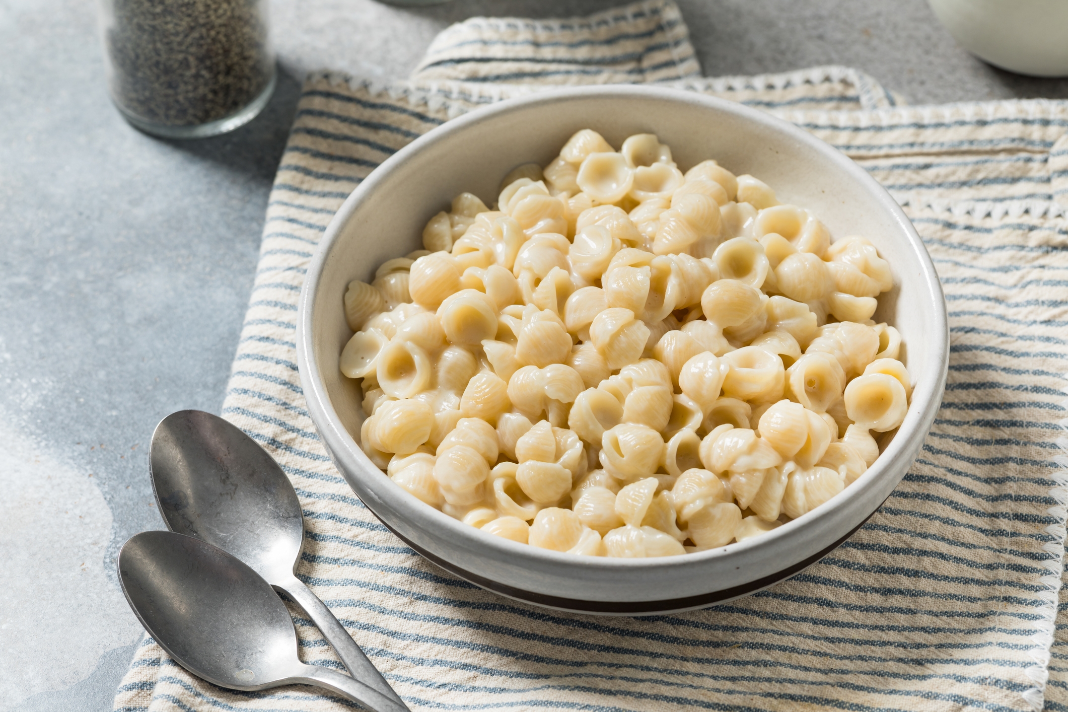A vessel of ammunition pasta with creamy food condiment connected a striped cloth, accompanied by 2 spoons and a capsicum shaker