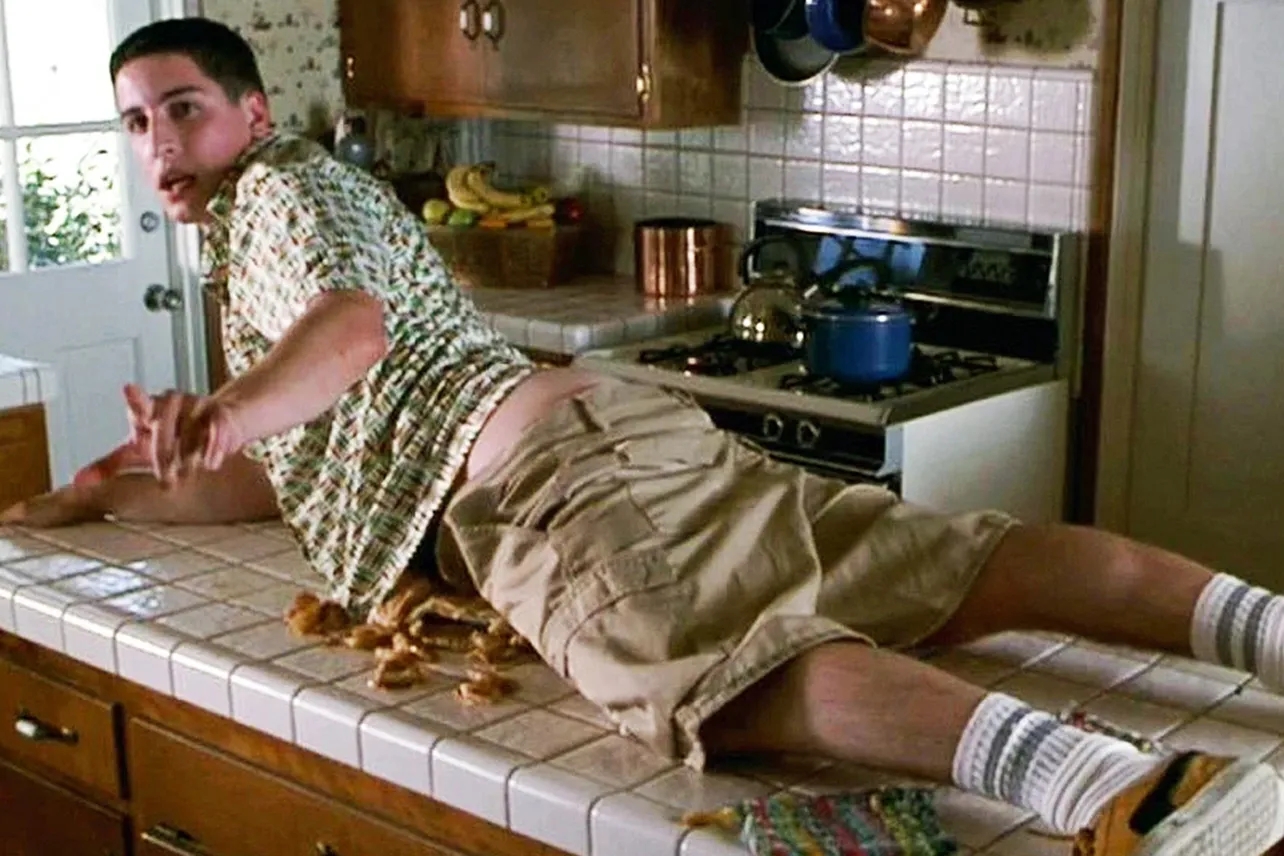 Person lying connected a room counter, surrounded by crumbs, wearing a patterned shirt, khaki shorts, and socks with sandals