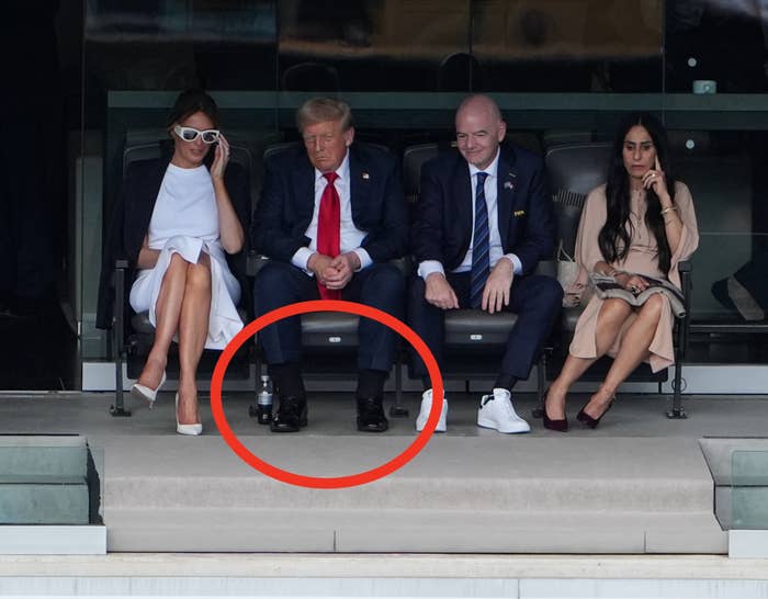 People seated in a stadium box, including a man in a suit and tie, watching the event below. Others are casually dressed or wearing formal attire