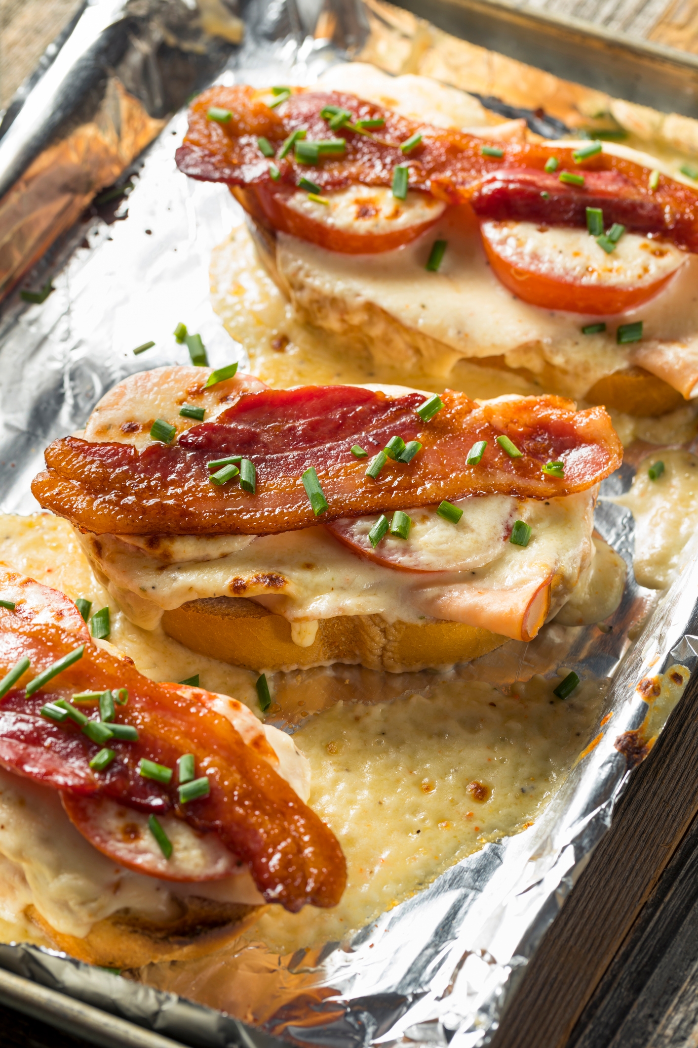 Baked sandwiches with melted cheese, tomato slices, bacon, and chives on a foil-lined tray