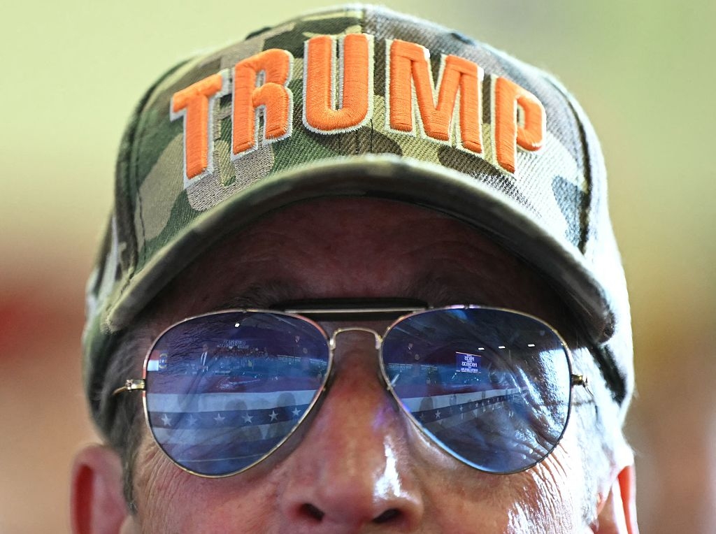 Person wearing a camouflage baseball cap with "TRUMP" embroidered, and mirrored sunglasses reflecting a flag pattern