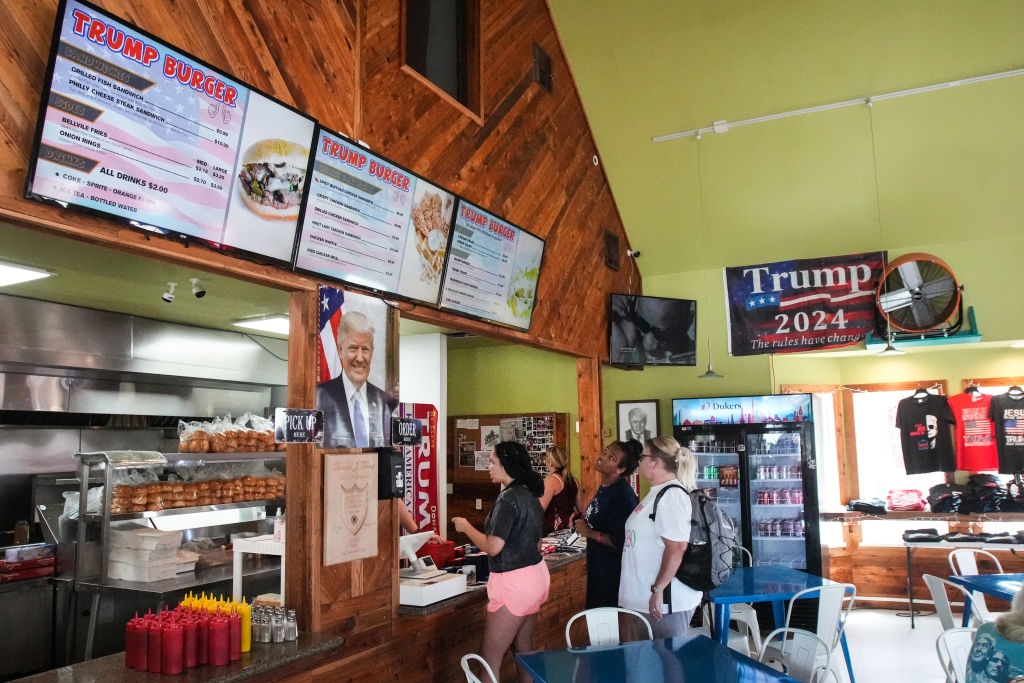 People ordering at a fast-food restaurant with "Trump 2024" merchandise visible and a menu above the counter displaying various burger options