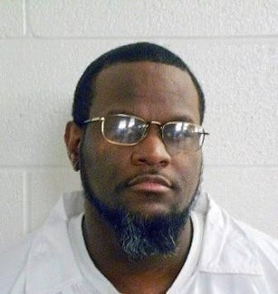 A person with glasses, a beard, and short hair against a plain wall, wearing a light shirt