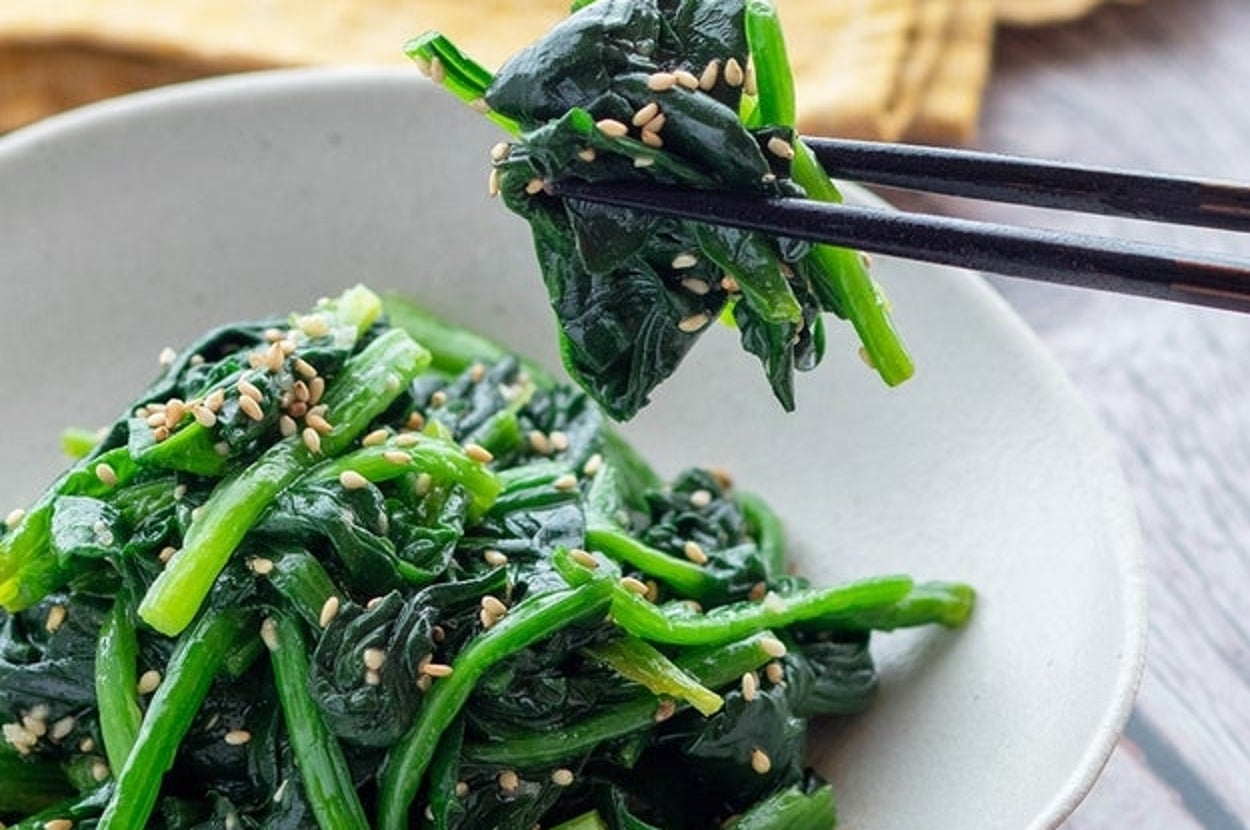 皿に盛られたほうれん草のおひたしを箸で持ち上げている様子。上にゴマがトッピングされている。