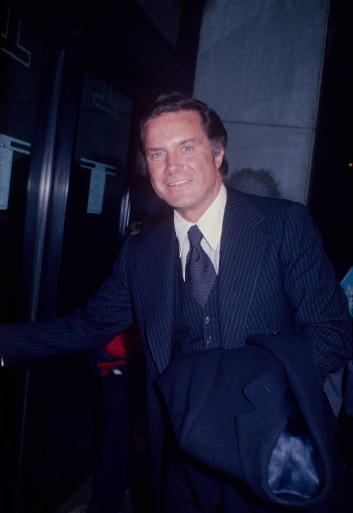 Cliff Robertson in a pinstripe suit holding a coat, smiling, standing near a glass door