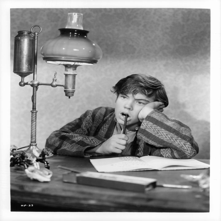 Bobby Driscoll in a patterned robe sits at a desk, appearing deep in thought, holding a pen with an open notebook in front