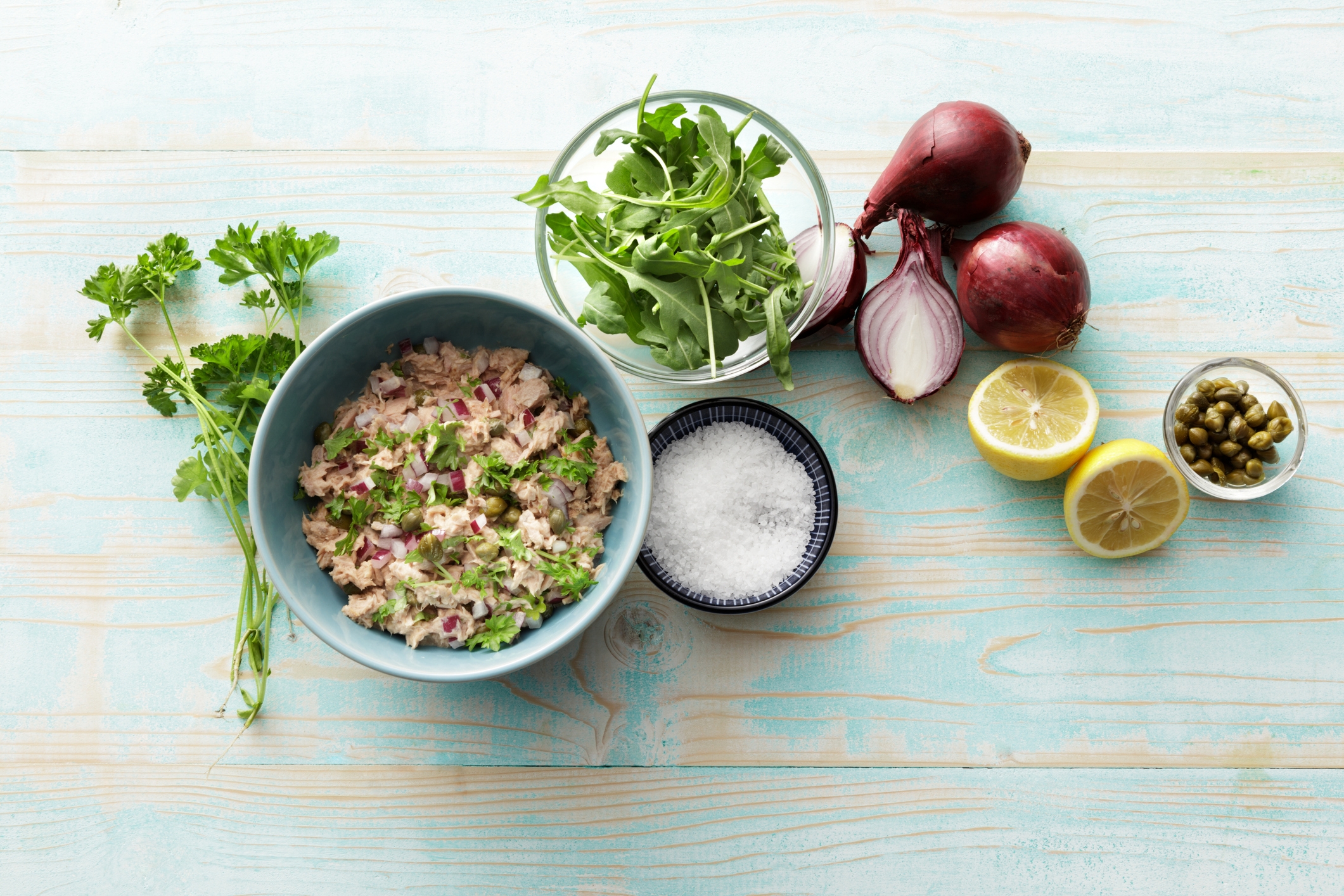  a vessel  of tuna salad, arugula, reddish  onions, citrus  halves, capers, parsley, and a brackish  dish