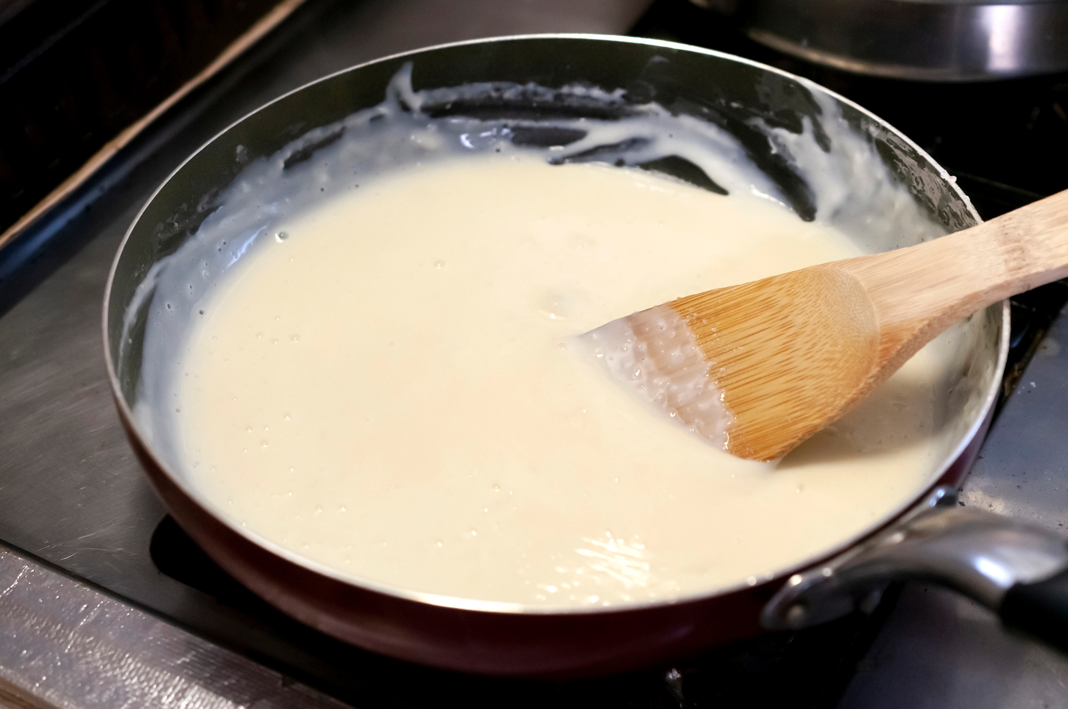 A saucepan with creamy achromatic  condiment  being stirred by a woody  spoon connected  a stovetop