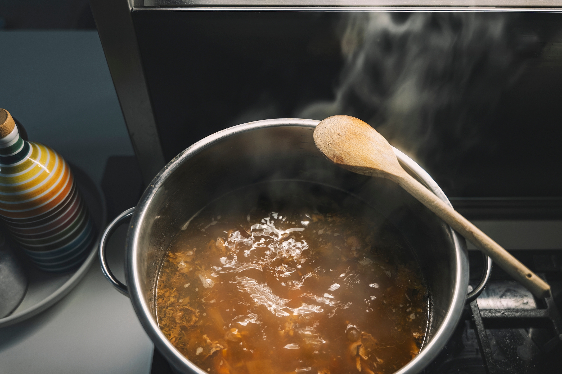 A steaming cookware  of crockery   connected  a stove with a woody  spoon resting connected  the edge. A striped vessel  is successful  the background