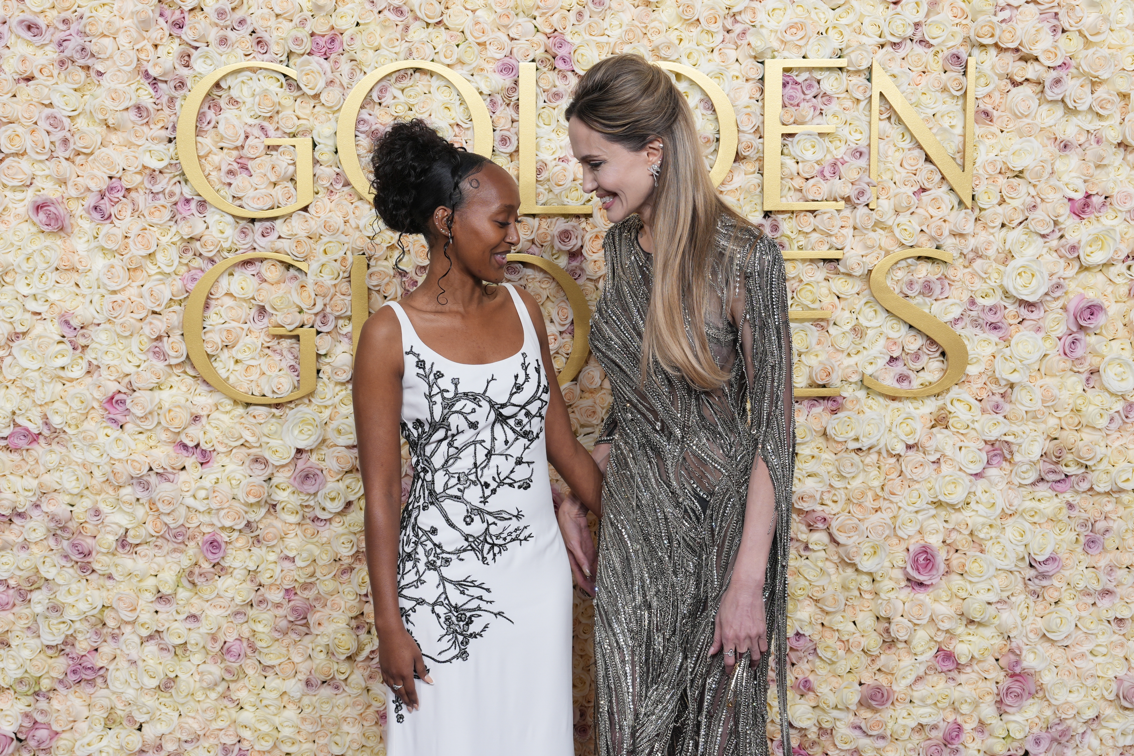 Two women basal successful beforehand of a floral backdrop. One wears a achromatic formal with achromatic detailing, the different a shimmering metallic gown