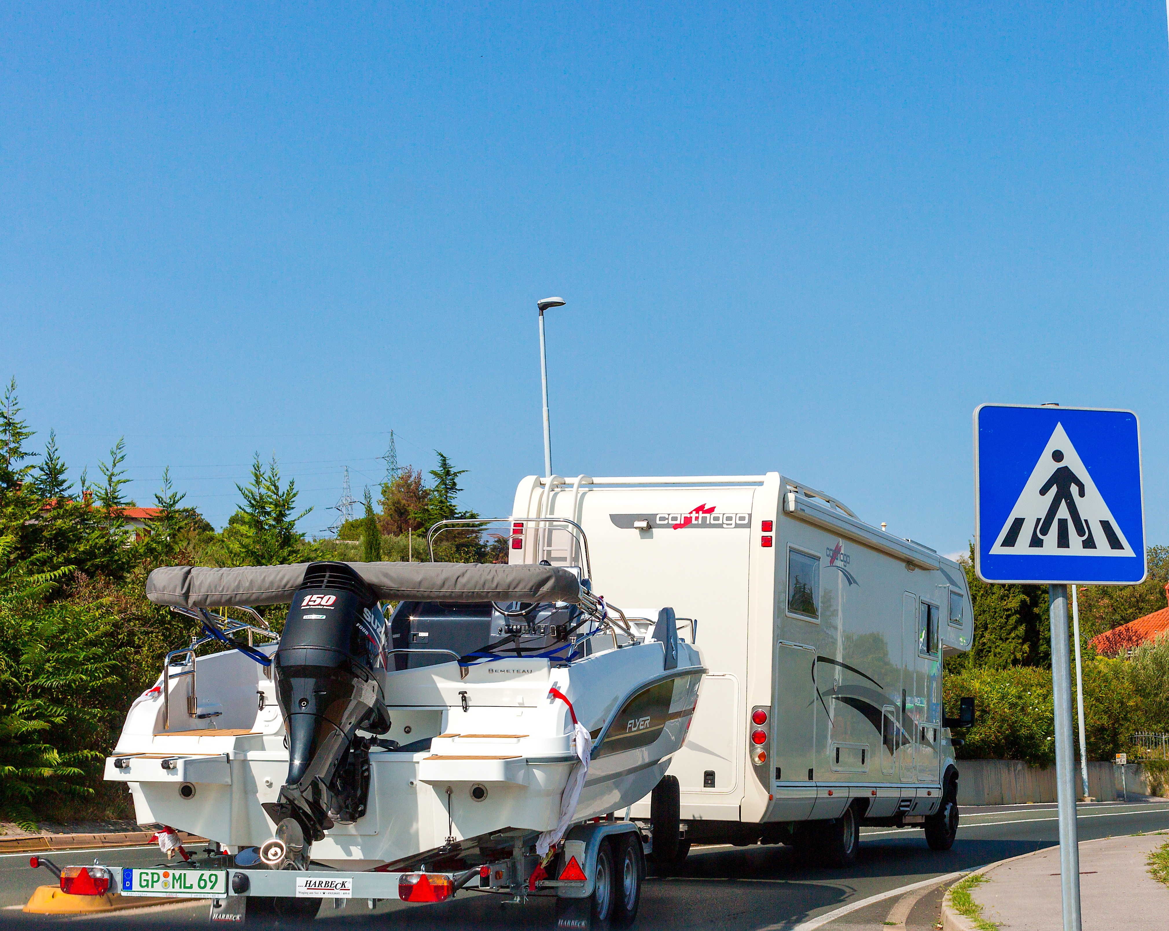 RV towing a vessel  connected  a roadworthy  adjacent   a pedestrian crossing sign, surrounded by trees and a wide   sky