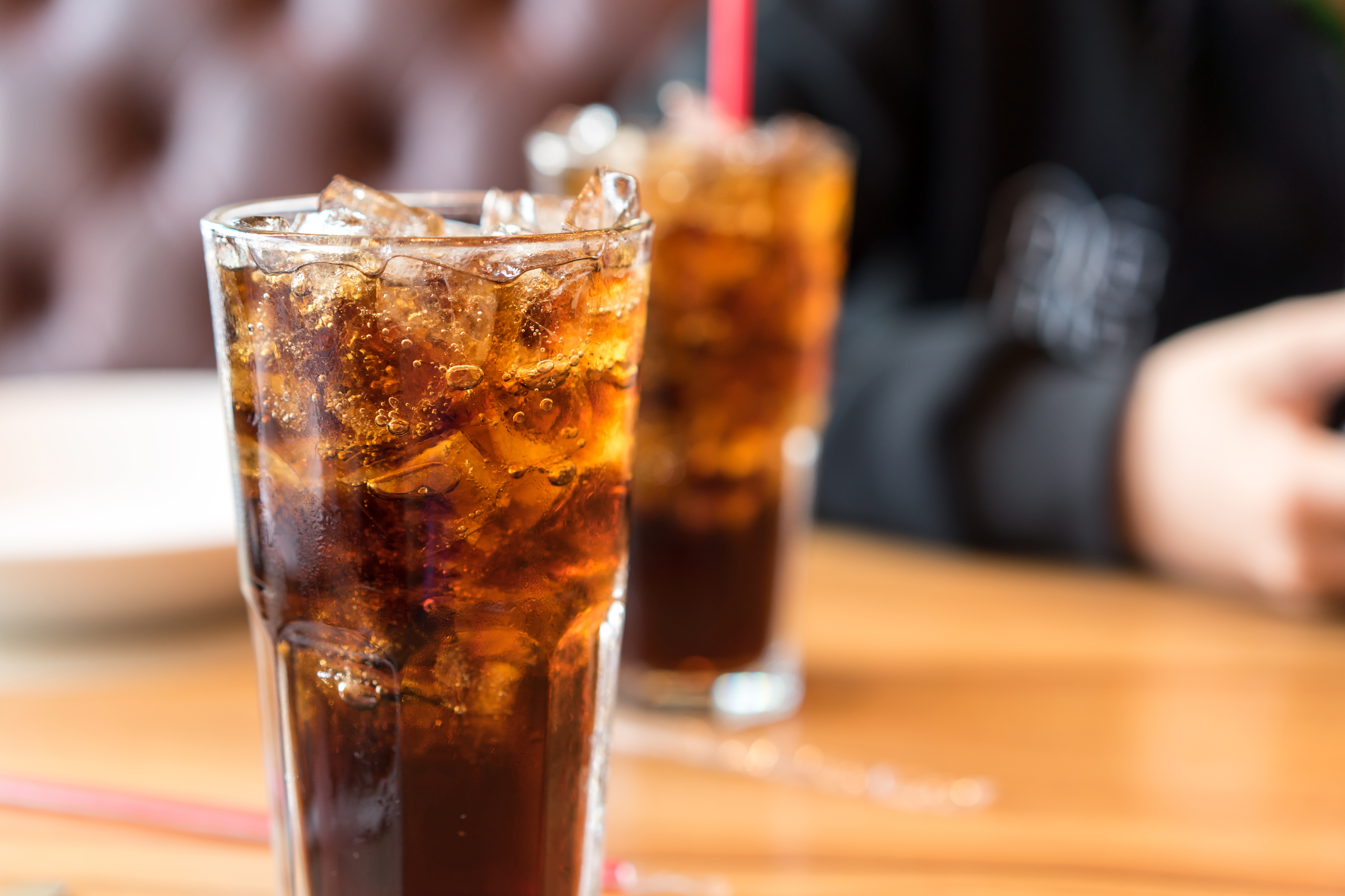 Two glasses of iced soda connected  a woody  table, with a blurred inheritance  suggesting a casual eating  setting
