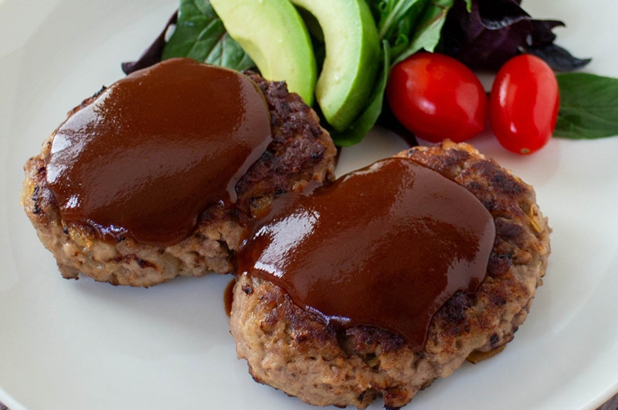 ハンバーグ2個がプレートに盛られ、アボカド、サラダ、プチトマトが添えられている。ソースがかかっている。