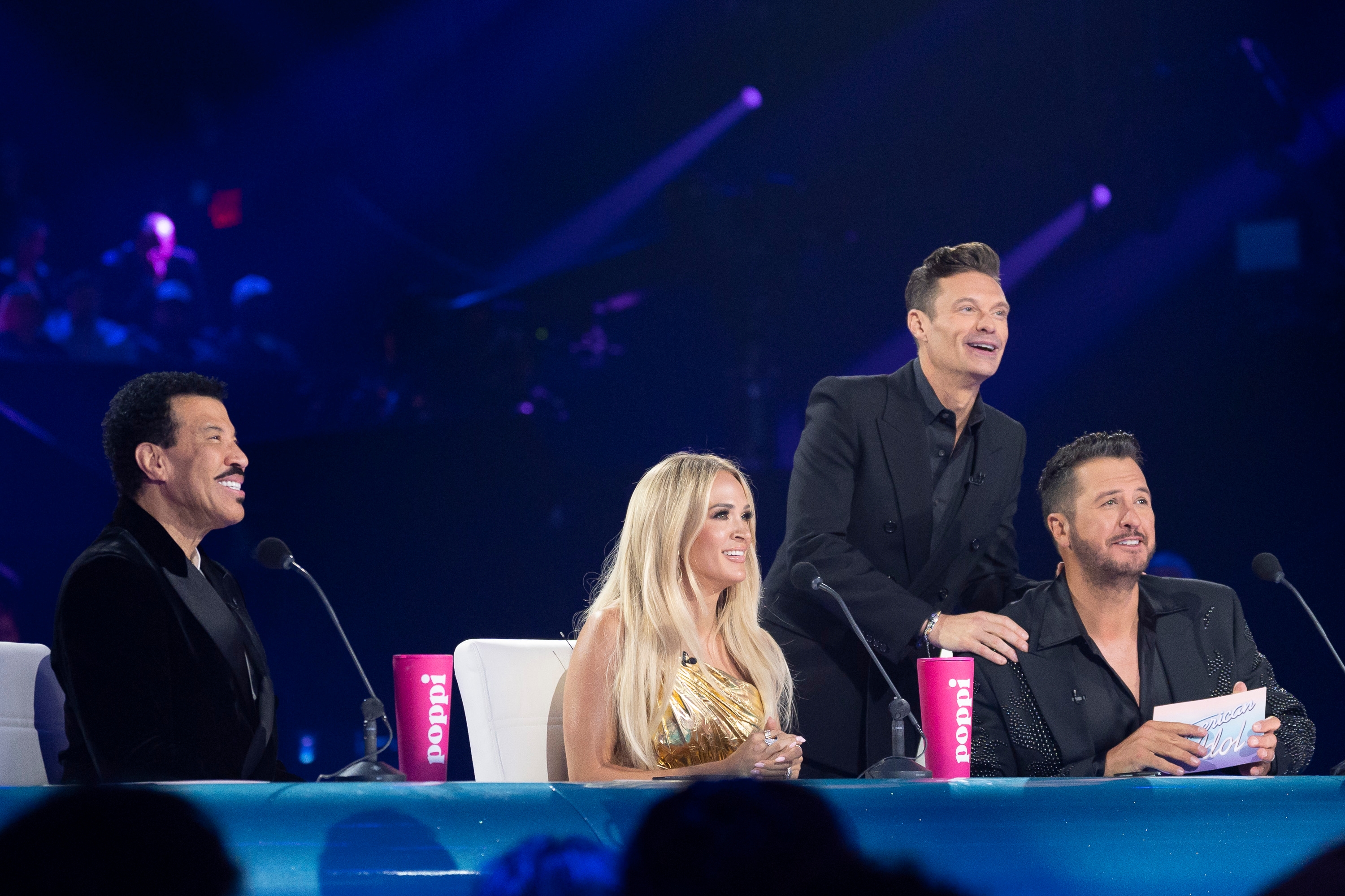 Lionel Richie, Carrie Underwood, and Luke Bryan dressed successful ceremonial attire, seated astatine a array with microphones with Ryan Seacrest lasting down Luke with a manus connected Luke's shoulder