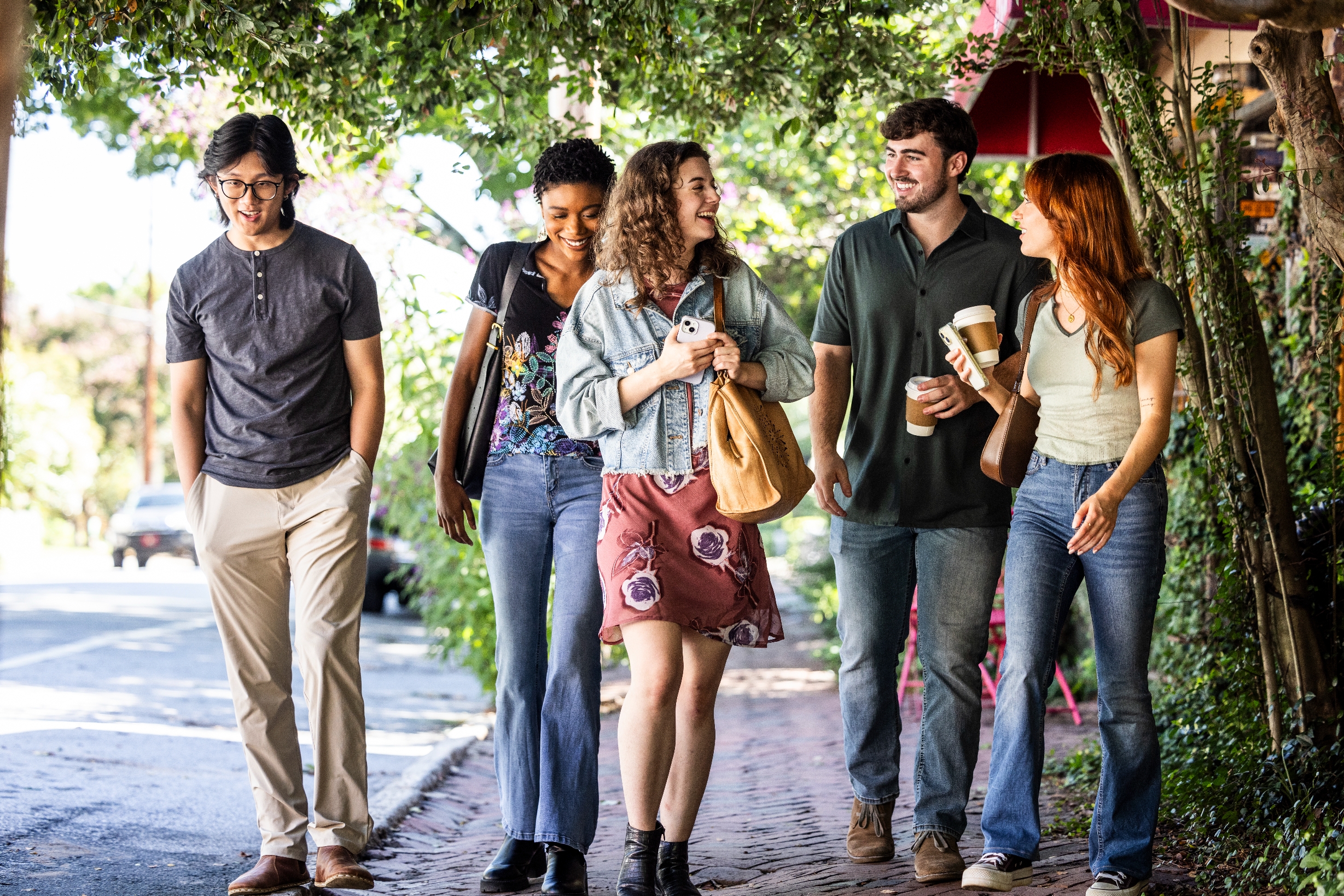 Group of 5  young adults walking connected  a sidewalk, laughing and chatting, each   holding java  cups, suggesting a casual outing oregon  break