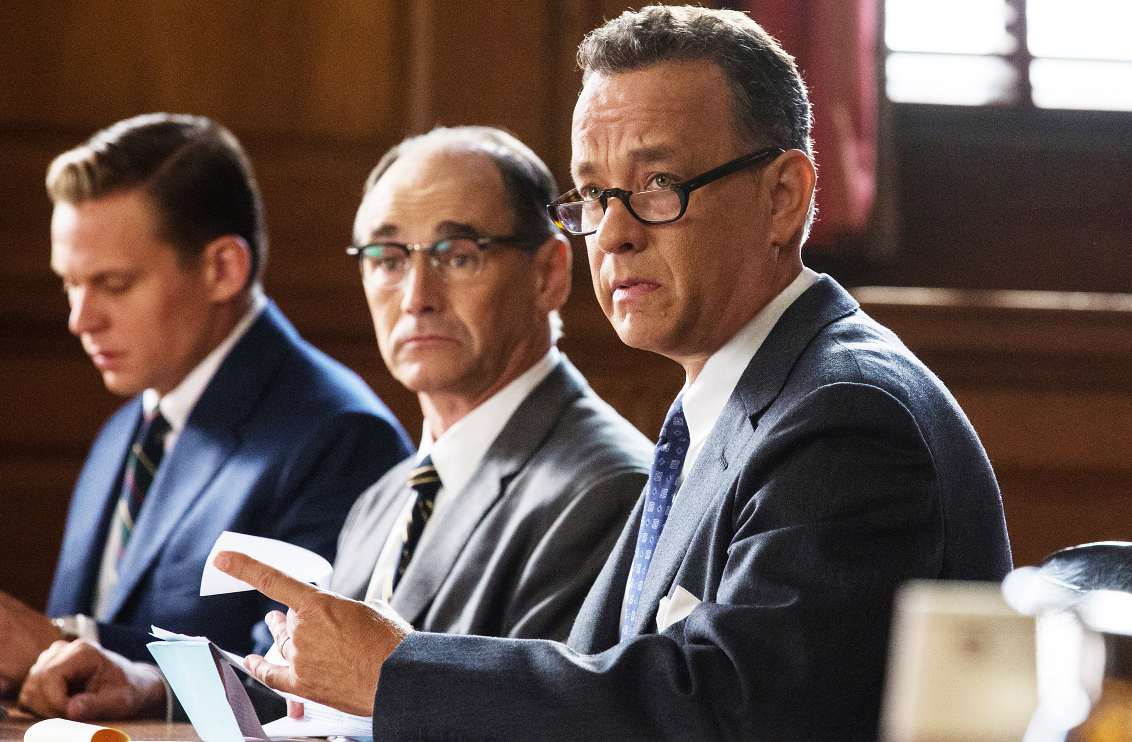 A man in a suit, with glasses, sitting at a table with two other men in suits, in what appears to be a courtroom setting