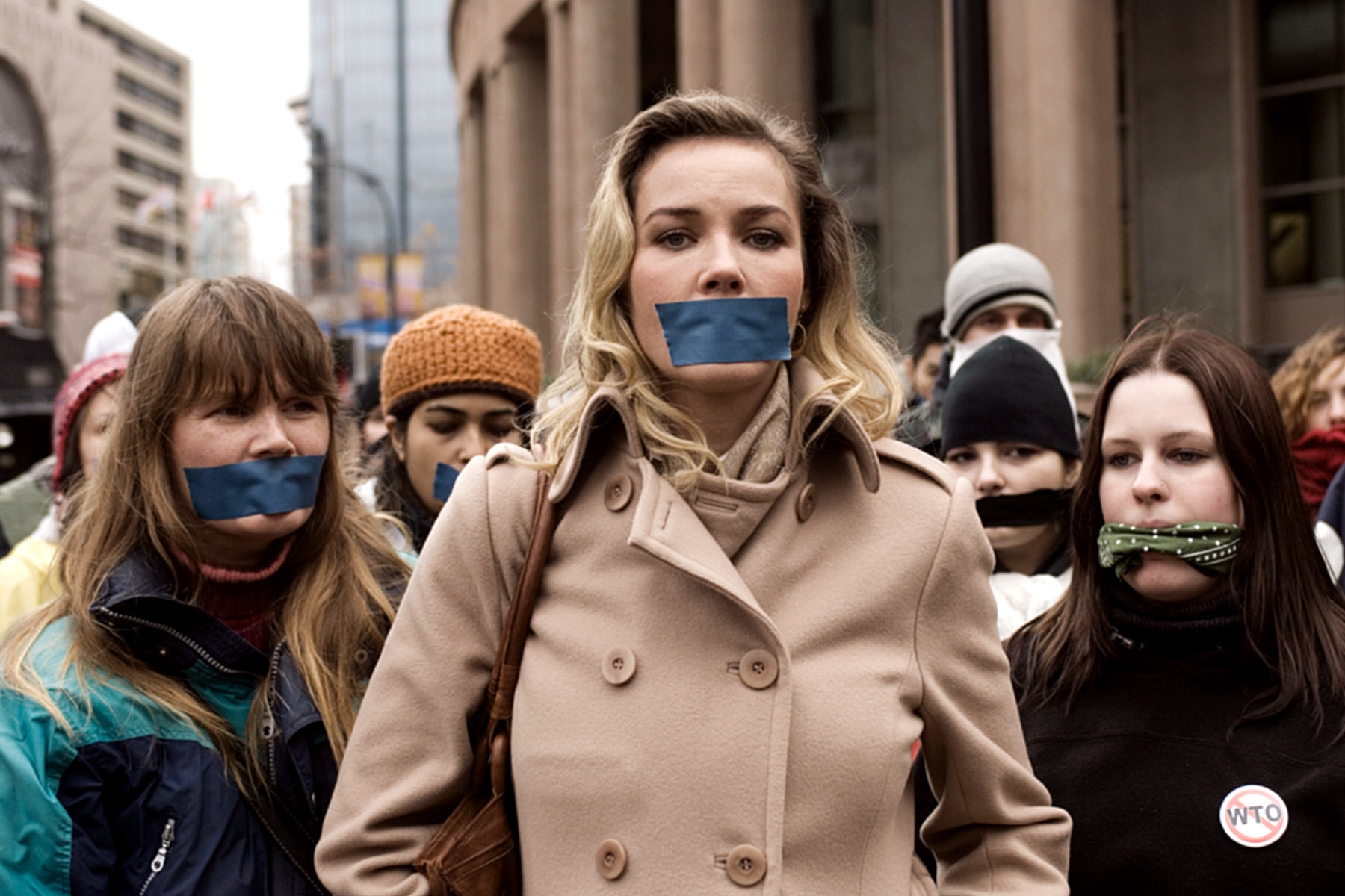 Group of protesters wearing coats stand with their mouths taped shut, symbolizing silenced speech. Urban setting in the background