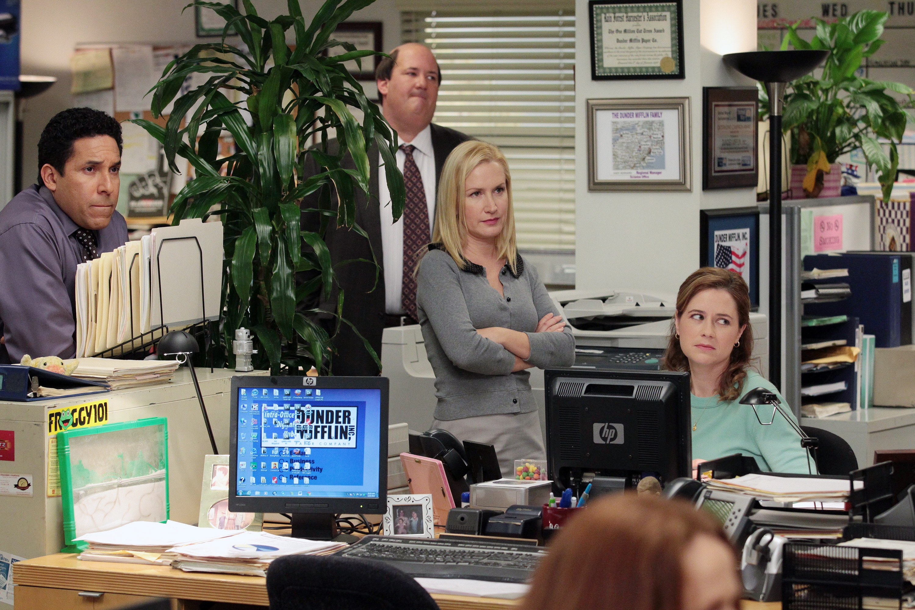 Four office workers gathered near desks in a fictional workplace. Computer screens visible with cluttered desks. Scene suggests casual interaction