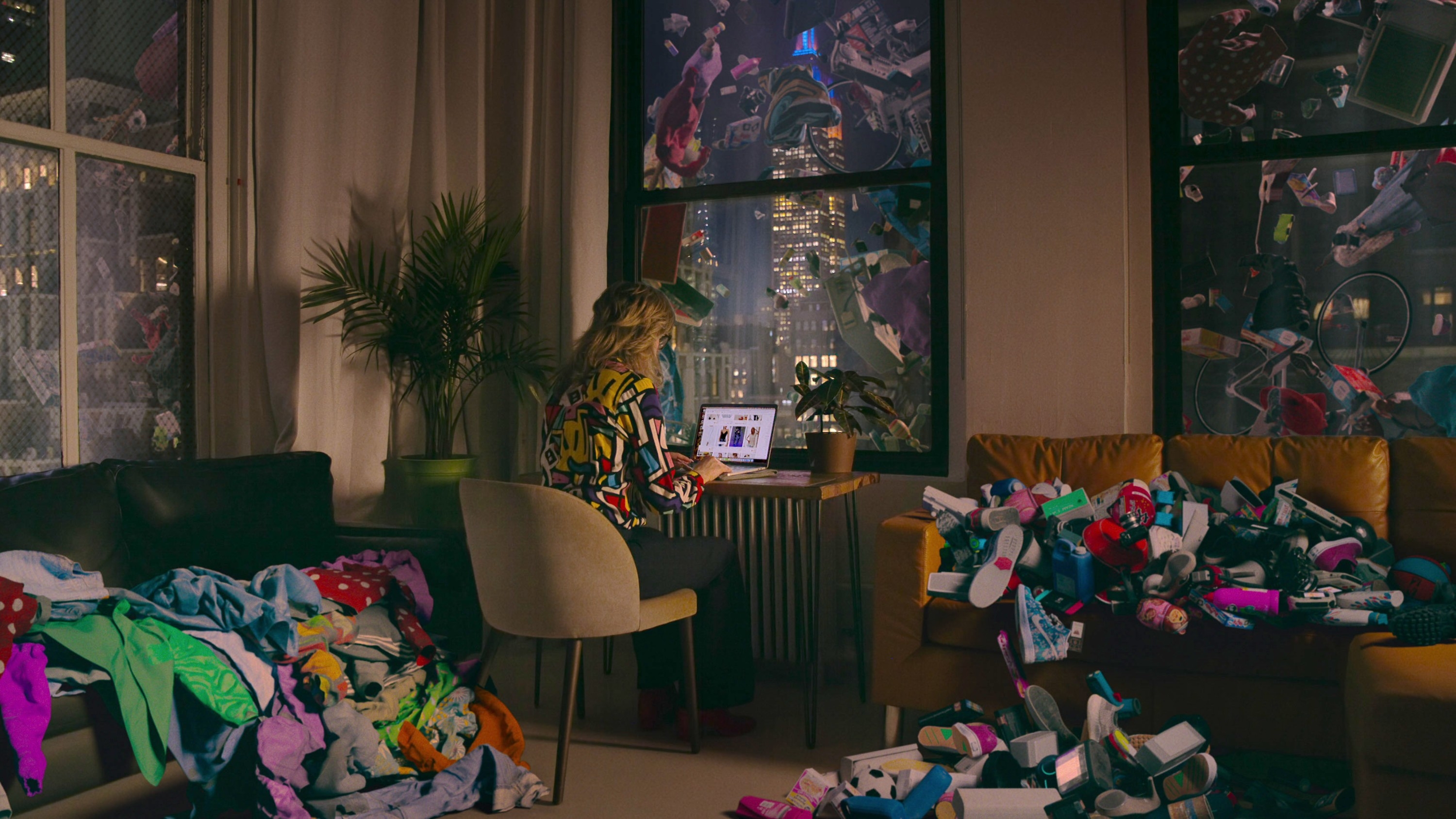 A person sits at a desk in a room cluttered with unfolded clothes while browsing a laptop, with city skyscrapers visible outside the windows