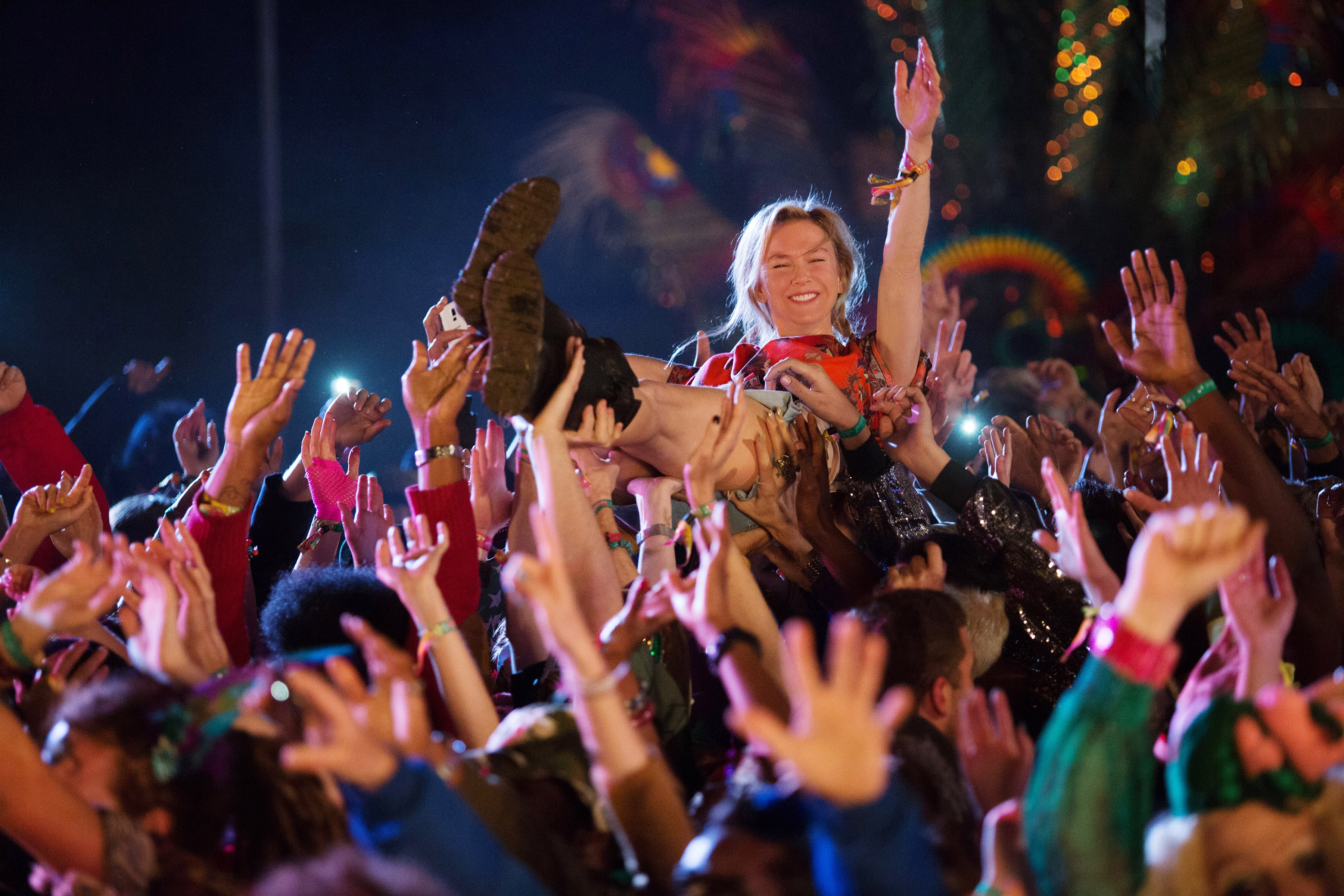 Person crowd surfing over a cheering audience, holding a shoe. Festival atmosphere with lights and energetic vibe