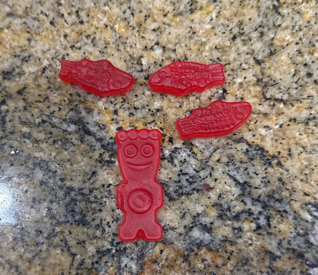 Four gummy candies shaped similar  food  and a smiling humanoid fig  are connected  a granite surface