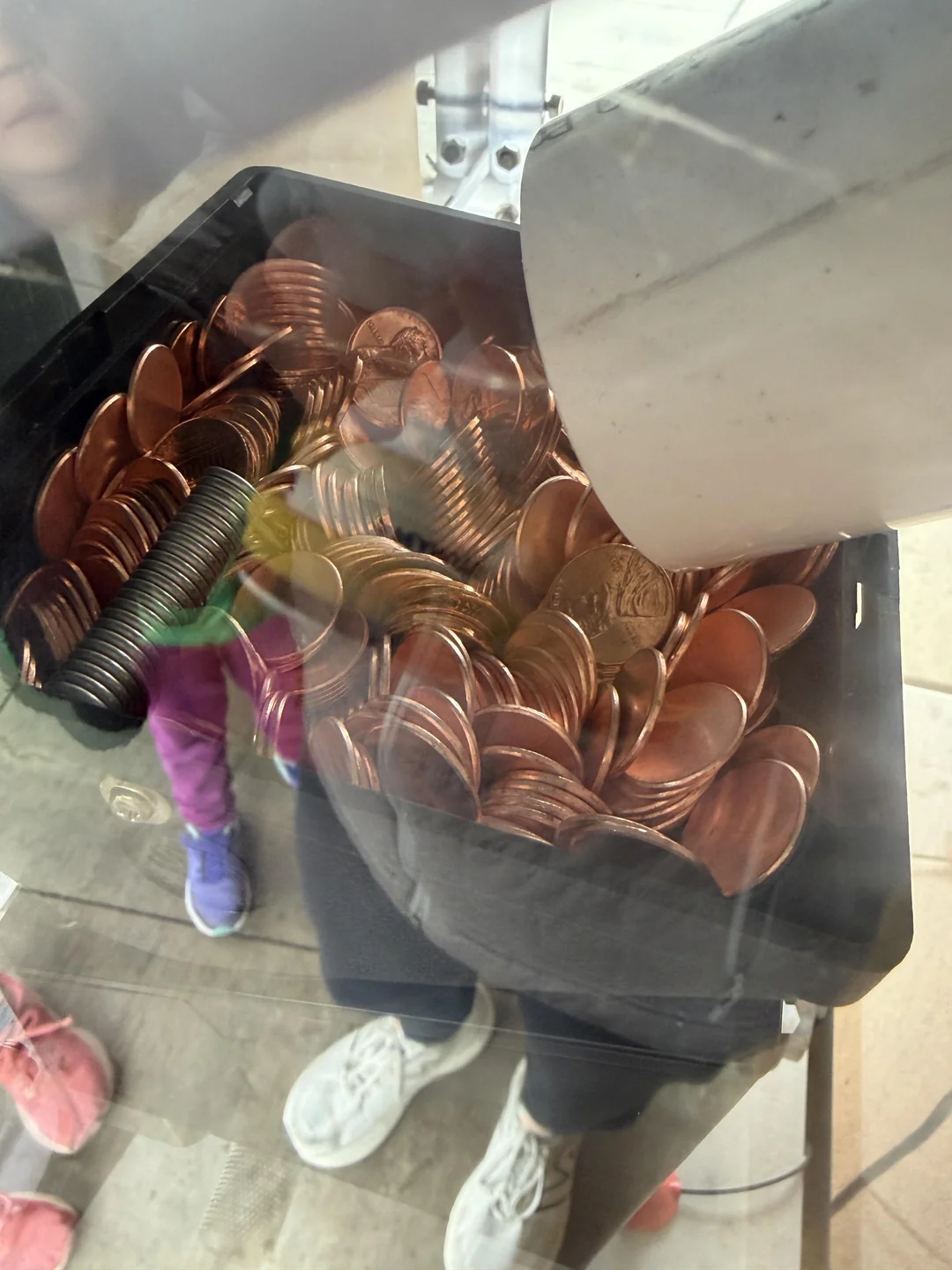 A coin sorter instrumentality   depositing pennies and dimes into a achromatic  tray. Reflections amusement   radical   lasting  nearby, wearing casual shoes