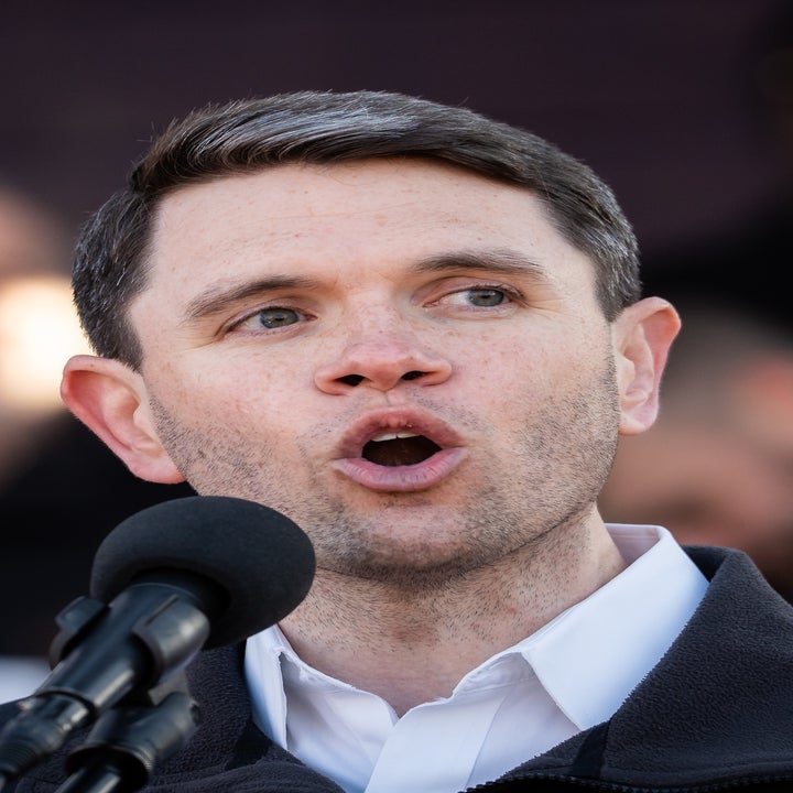 A person speaking passionately into a microphone at an outdoor event with a blurred crowd in the background
