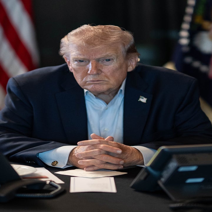 A man in a suit sits at a desk with phones and papers, looking serious, with flags in the background