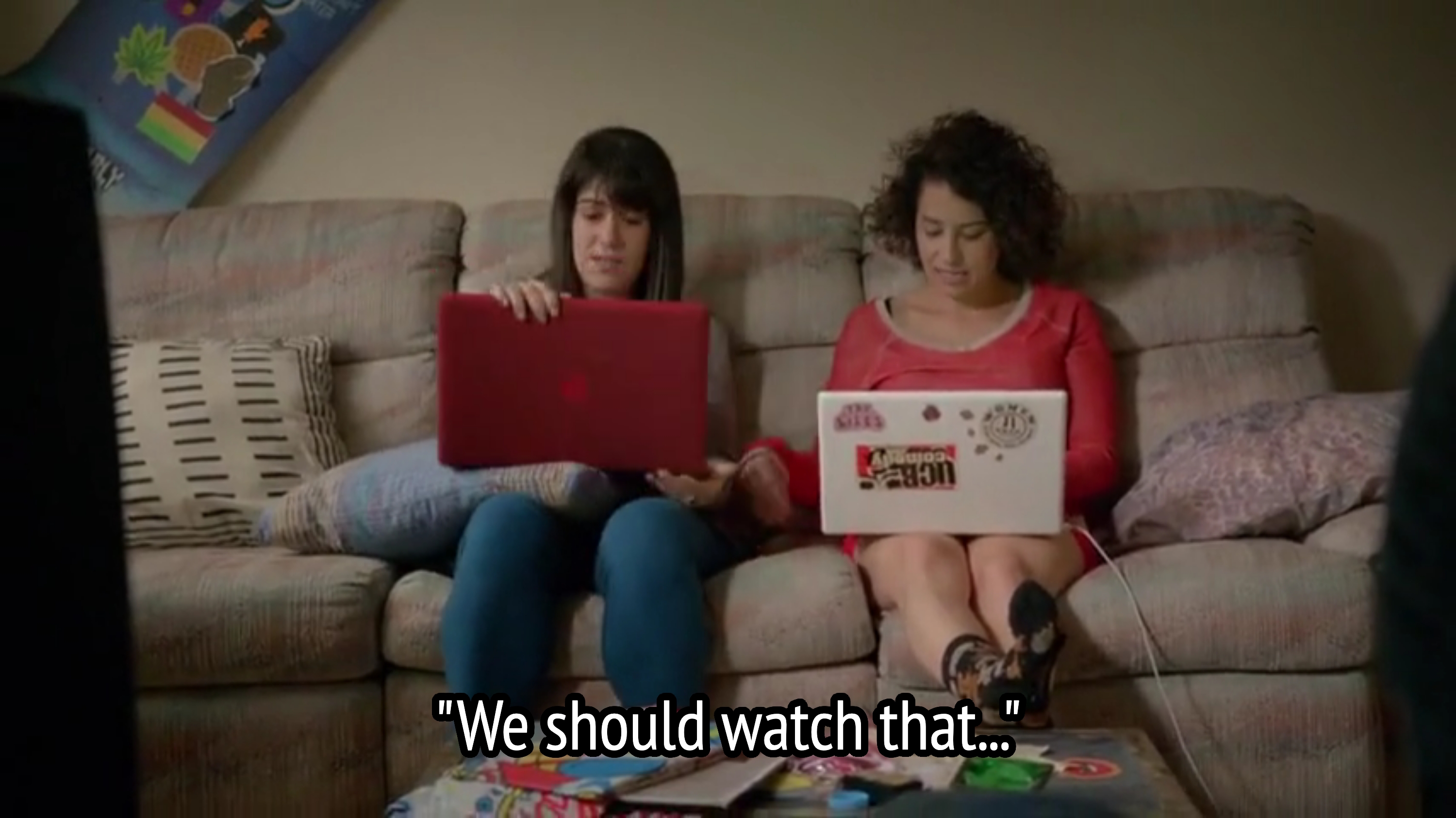 Two radical   sitting connected  a couch, each   utilizing a laptop. One has a reddish  laptop; the different   has a achromatic  laptop with stickers. A array  with items is successful  front