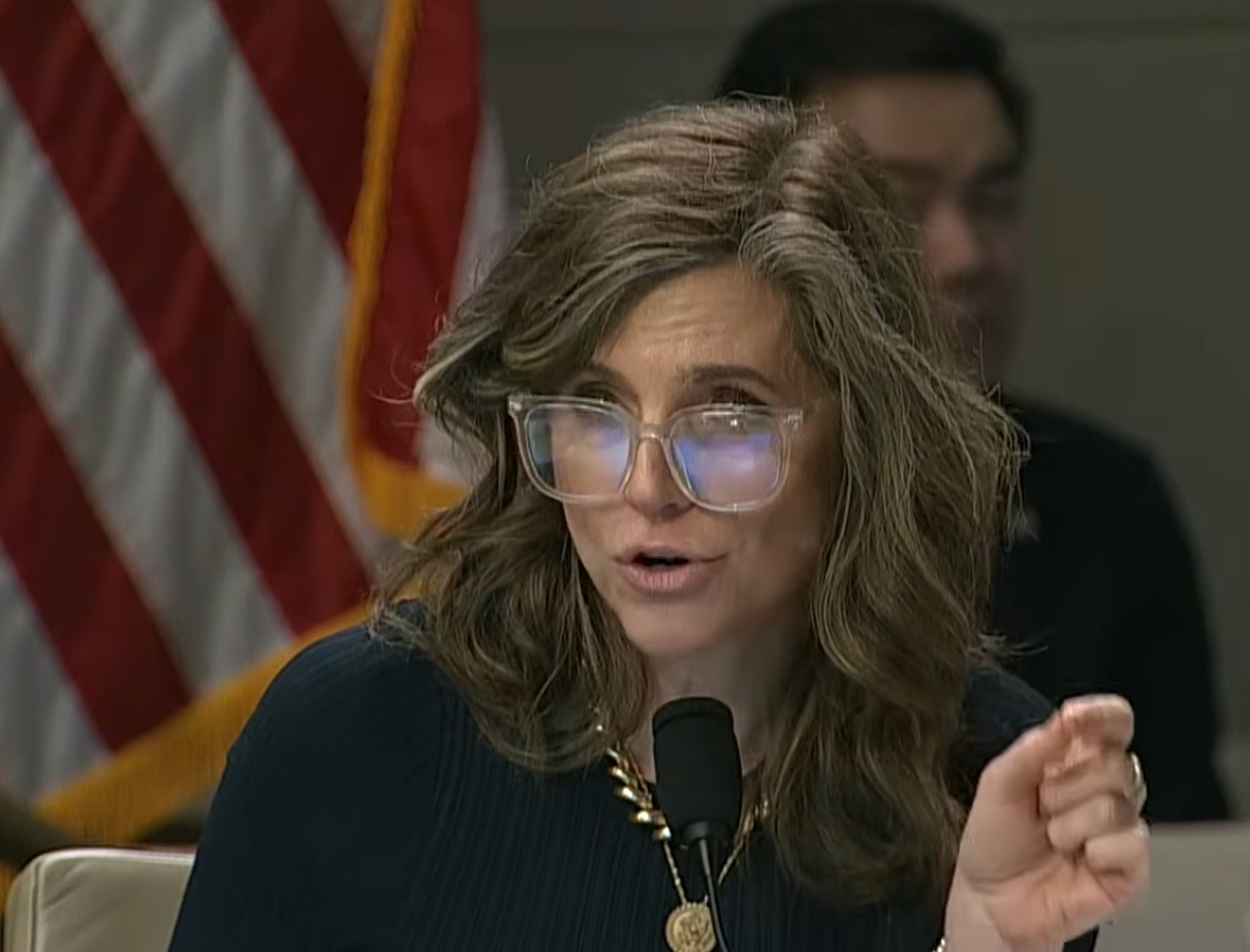 Person with agelong  hairsbreadth  and glasses speaks into a microphone during a meeting, an American emblem  disposable   successful  the background