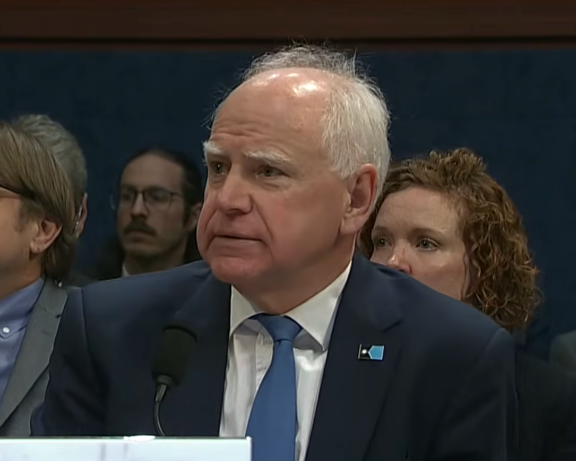 An older antheral   successful  a suit   testifying astatine  a hearing. People are seated down  him, showing interest