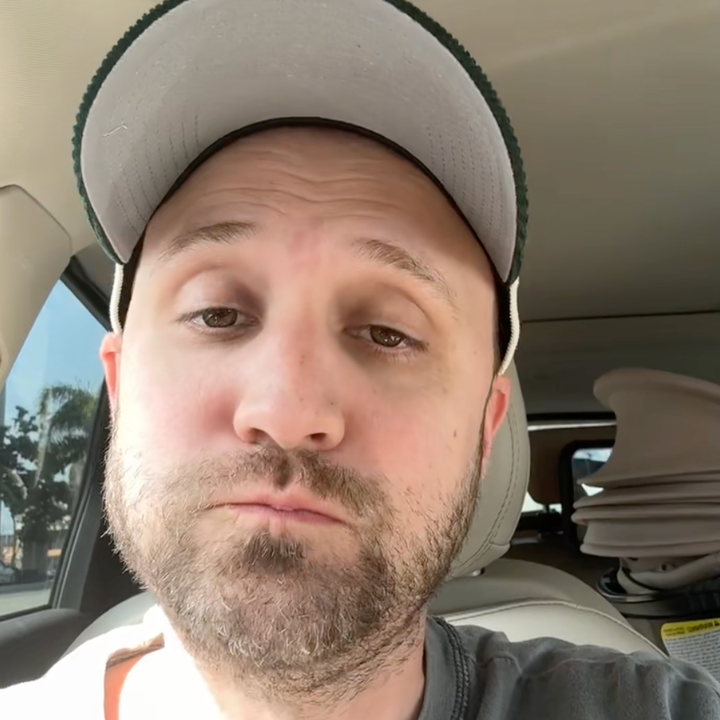 A person with a neutral expression sits in a car, wearing a cap and a casual shirt. A car seat is visible in the background