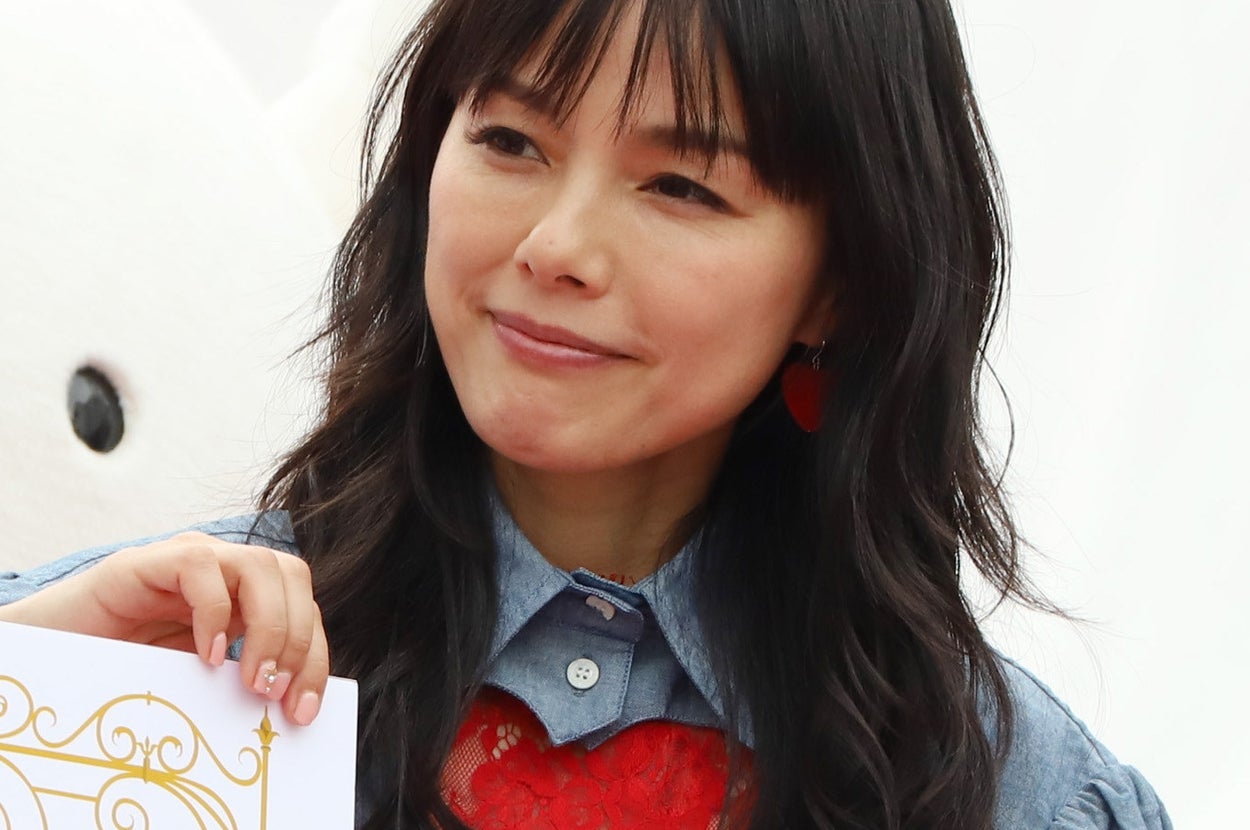 女性が赤いハートの柄が特徴的な青いシャツを着て微笑む。手に装飾された紙を持っている。