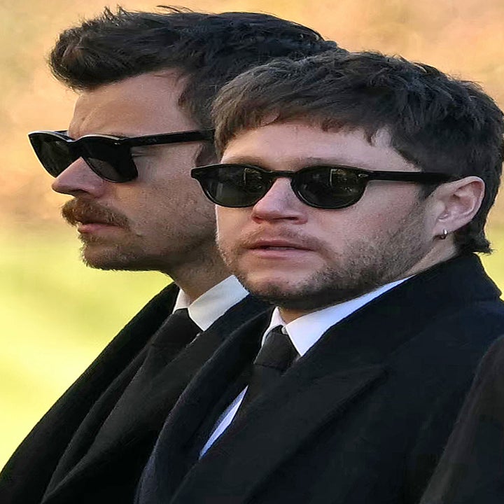 Two men in suits and sunglasses are standing side by side, looking in the same direction