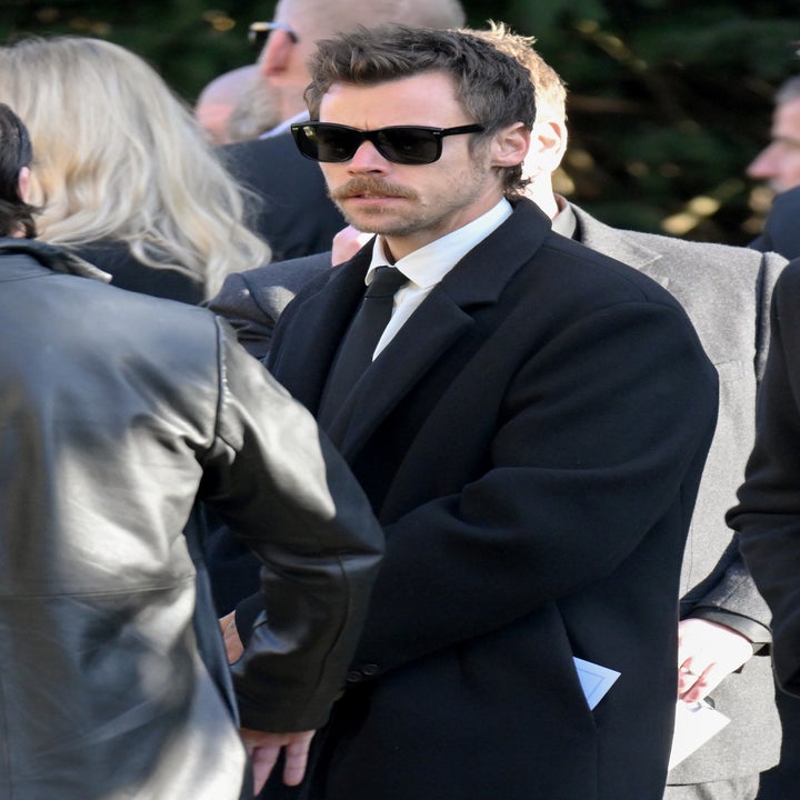 Man in a suit with sunglasses and a mustache stands outdoors among others, holding a folded paper