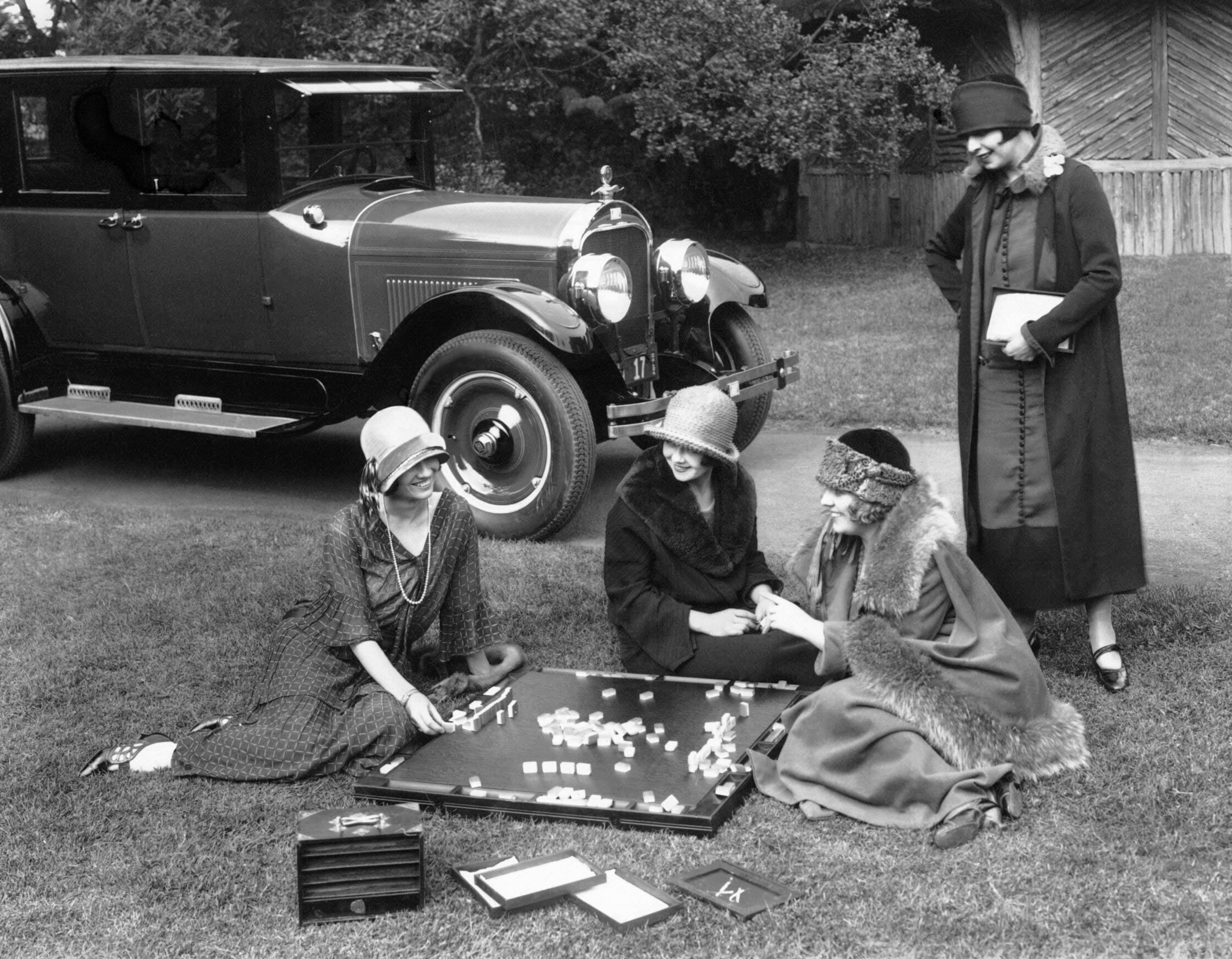 Four women successful  1920s attire play   a crippled  connected  the writer  beside a vintage car, exuding an elegant, nostalgic vibe