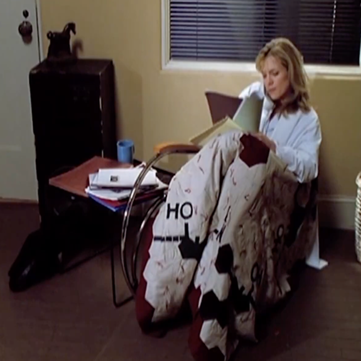 A person sits on the floor, wrapped in a patterned blanket, reading a book next to a small table with papers and a coffee cup