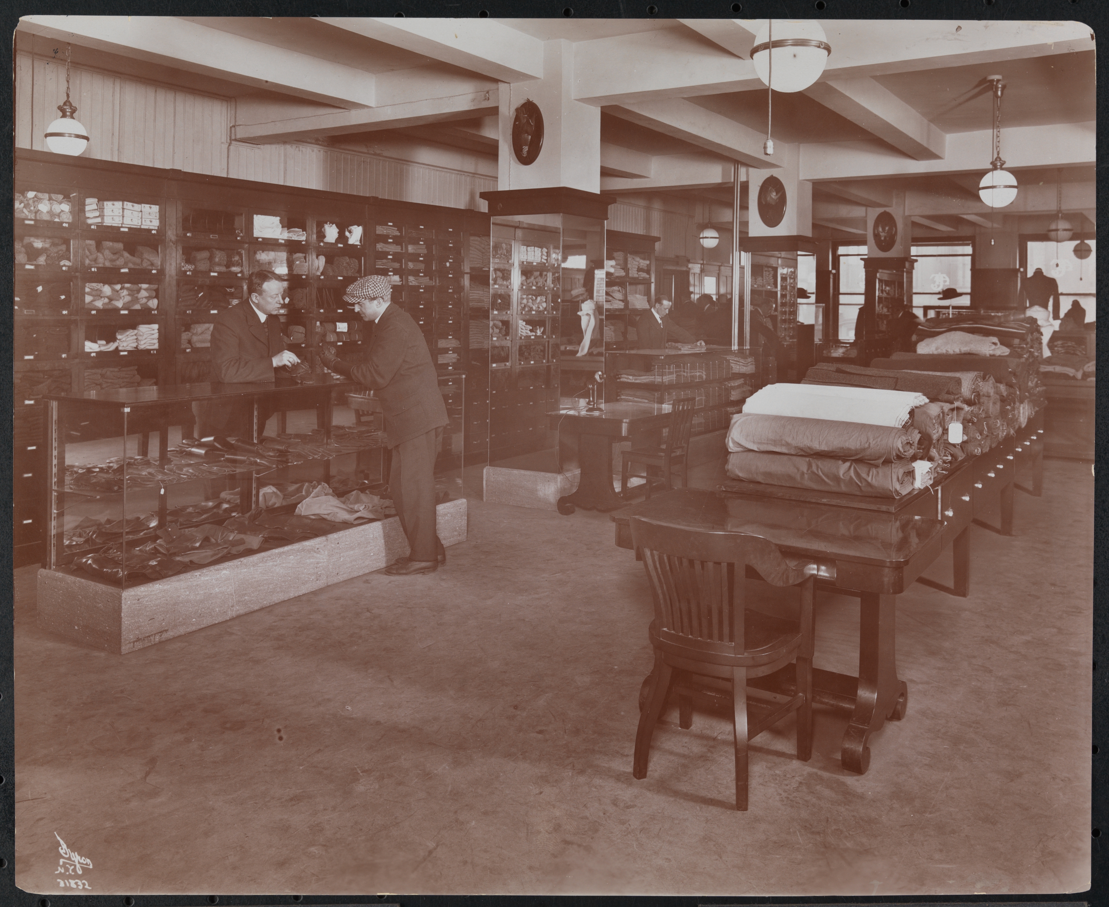Vintage store   interior with men successful  suits interacting adjacent   shelves filled with goods. Tables show  fabrics. Early 20th-century buying  scene