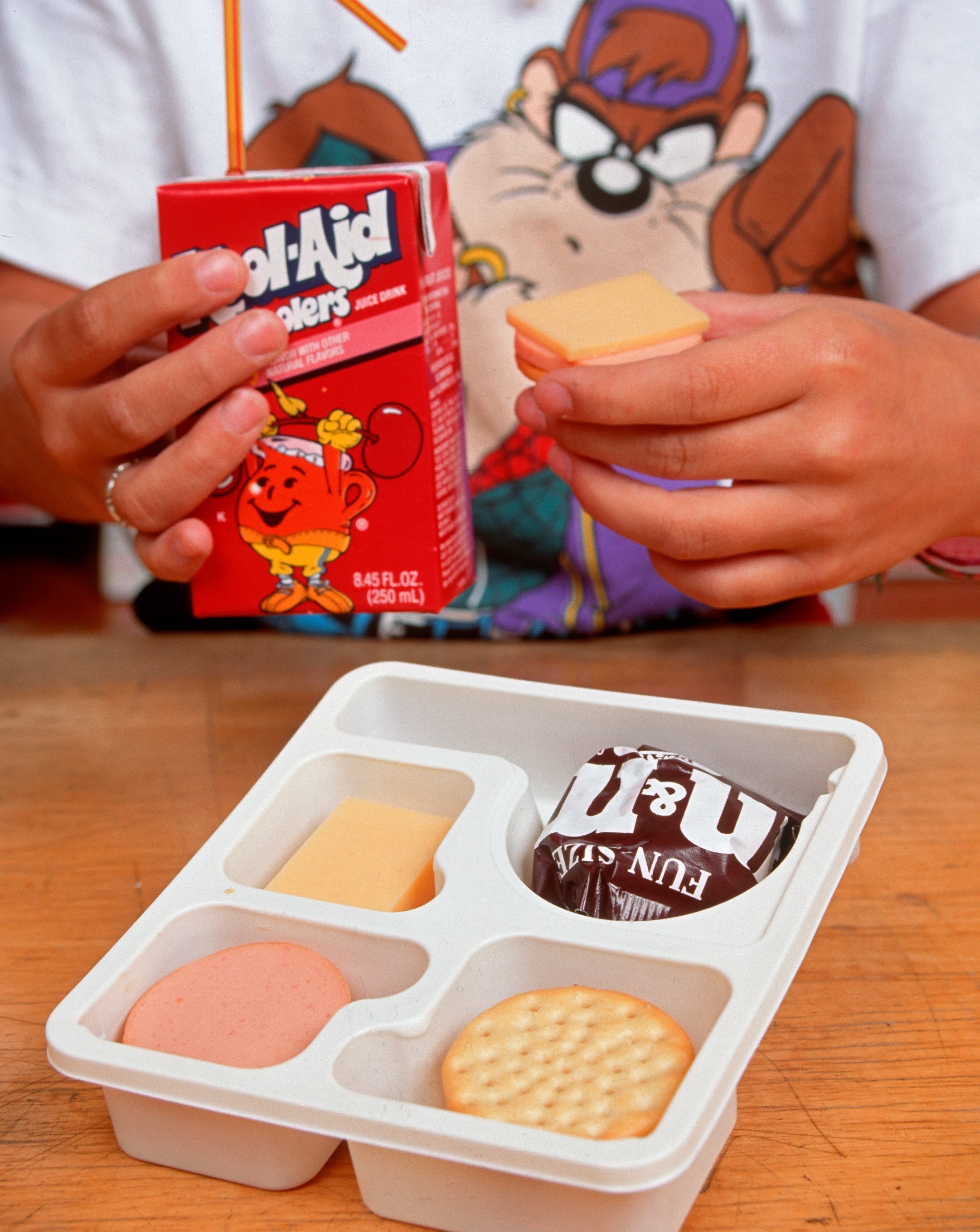 Person holding a Kool-Aid Jammers portion  adjacent  to a Lunchables tray with crackers, cheese, and meat. They're wearing a T-shirt with a cartoon character