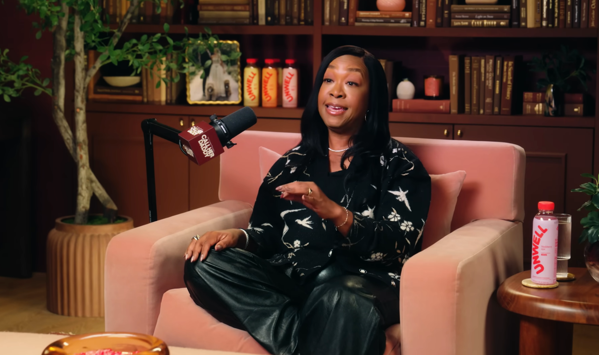 Person sitting connected  a sofa  successful  a studio, speaking into a microphone. They are wearing chic floral attire and surrounded by bookshelves and decor